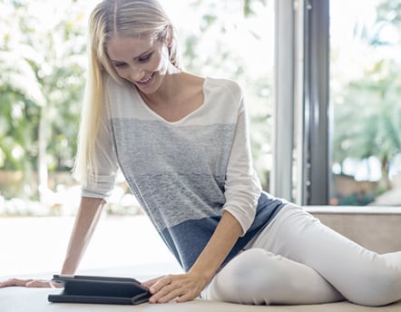 Eine Frau mit langen blonden Haaren, die einen hellen Pullover und eine weiße Hose trägt, sitzt drinnen an einem großen Fenster und schaut lächelnd auf ein Tablet, das vor ihr auf dem Tisch liegt.
