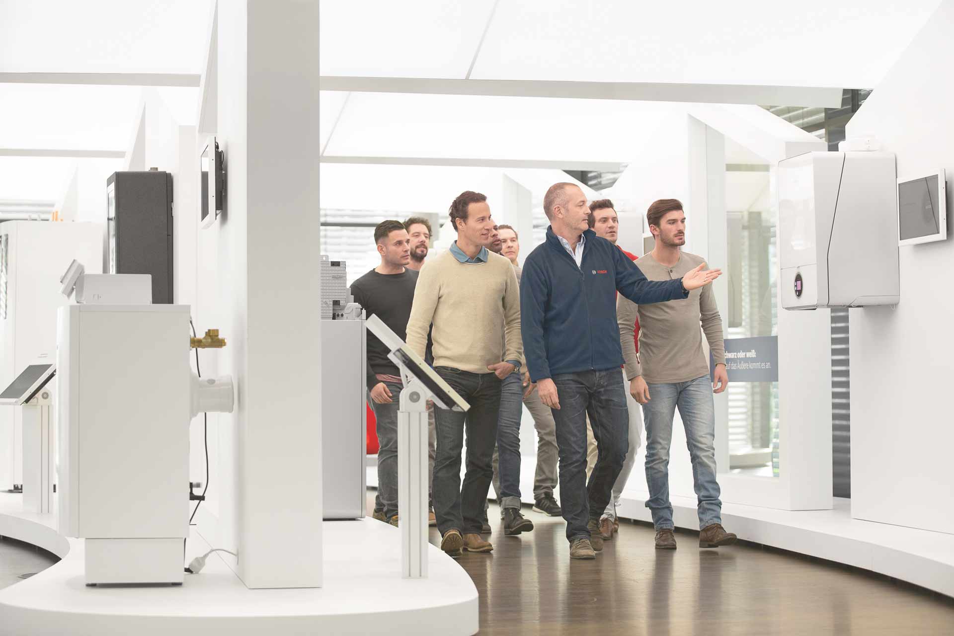 Eine Gruppe von Männern geht durch einen modernen Ausstellungsraum mit weißen Wänden und Displays, angeführt von einem Mann in einer blauen Jacke, der etwas zu erklären scheint.