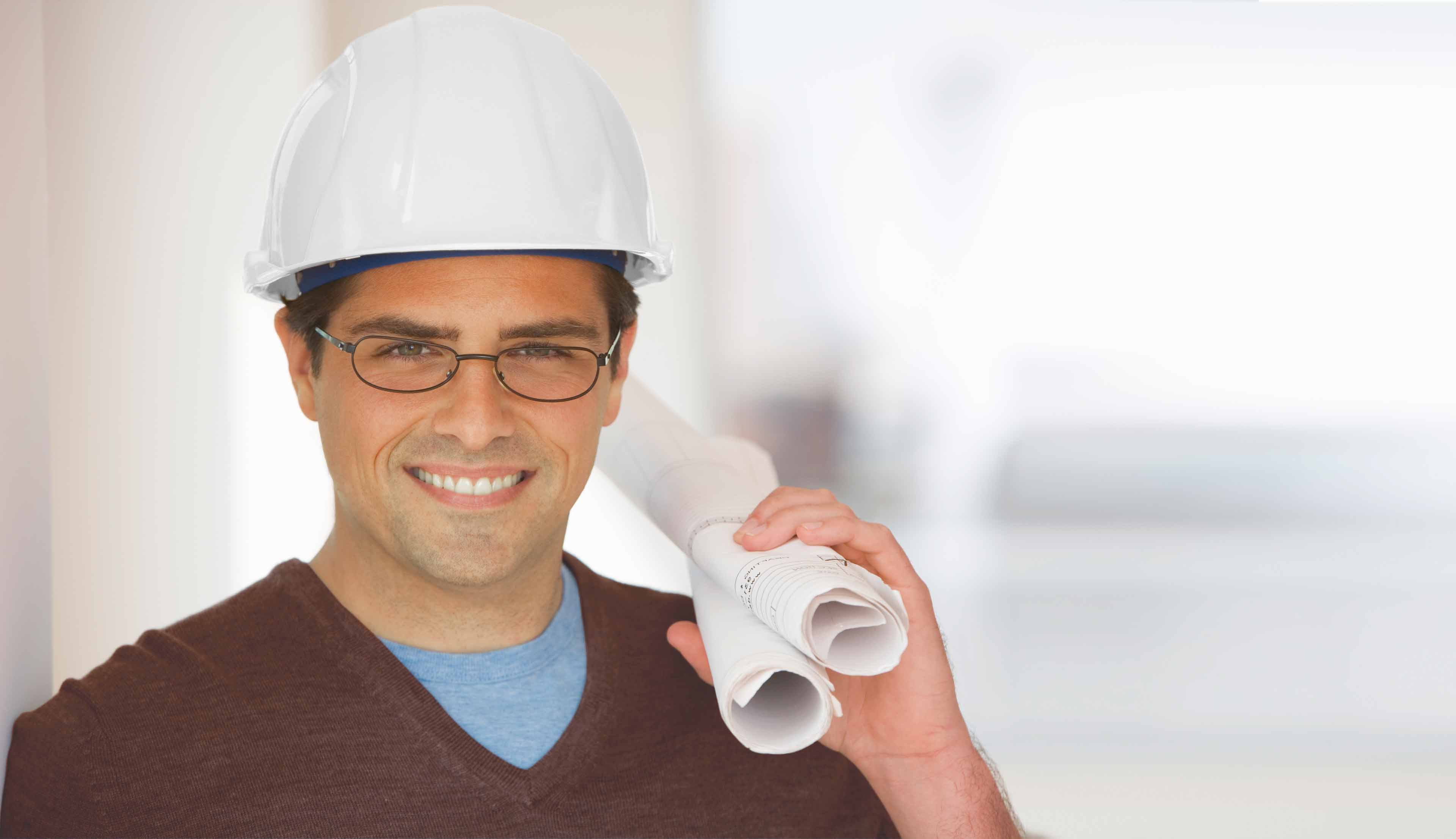 Ein Mann, der eine Brille und einen weißen Bauhelm trägt, lächelt und hält aufgerollte Baupläne über seine Schulter. Er ist mit einem braunen Hemd mit V-Ausschnitt bekleidet und steht in einem hellen Innenbereich.
