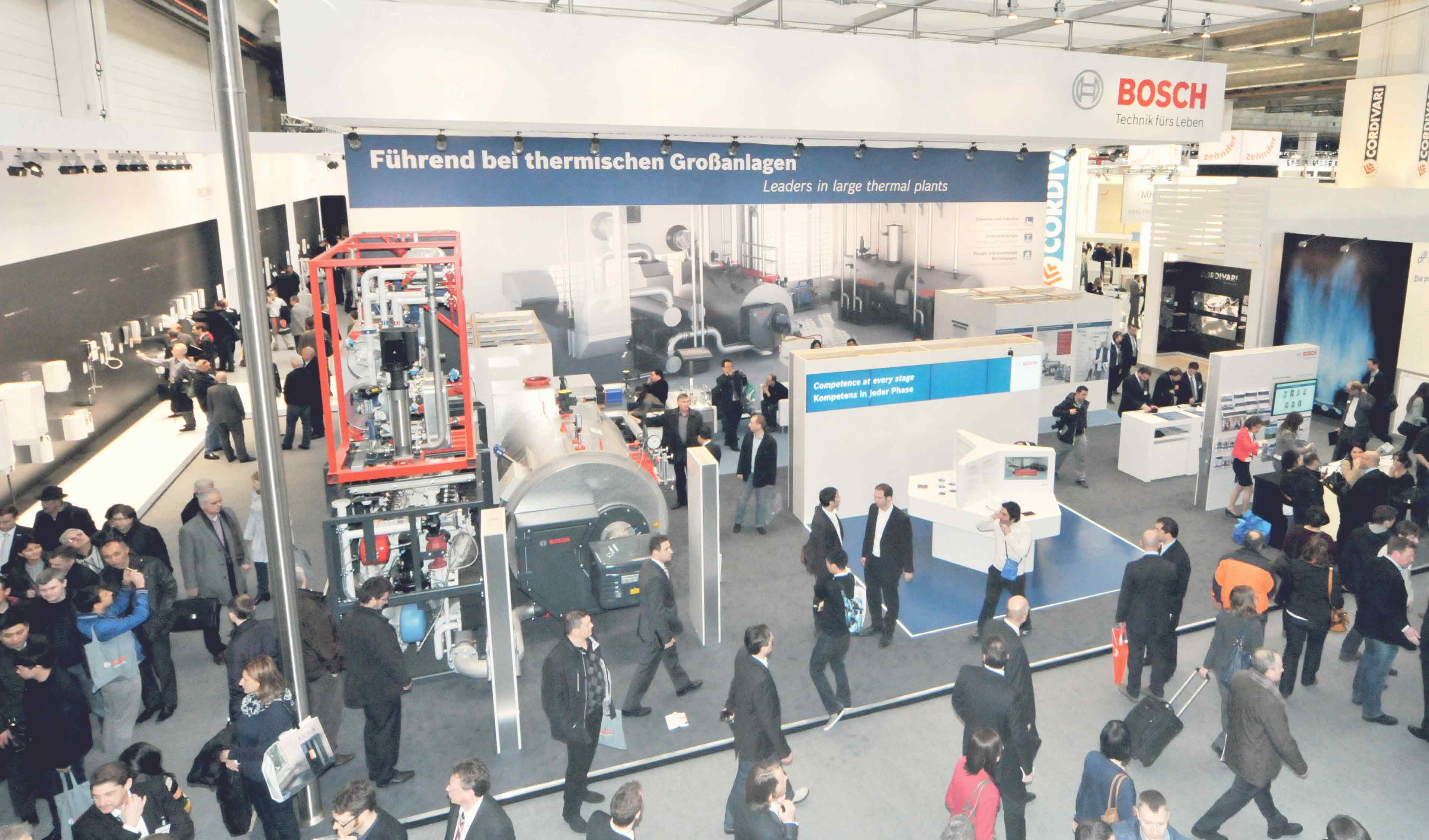 Eine belebte Messehalle mit großen Industrieausrüstungen, vielen Ständen und zahlreichen Menschen, die herumlaufen; ein Bosch-Schild und ein Banner mit der Aufschrift 