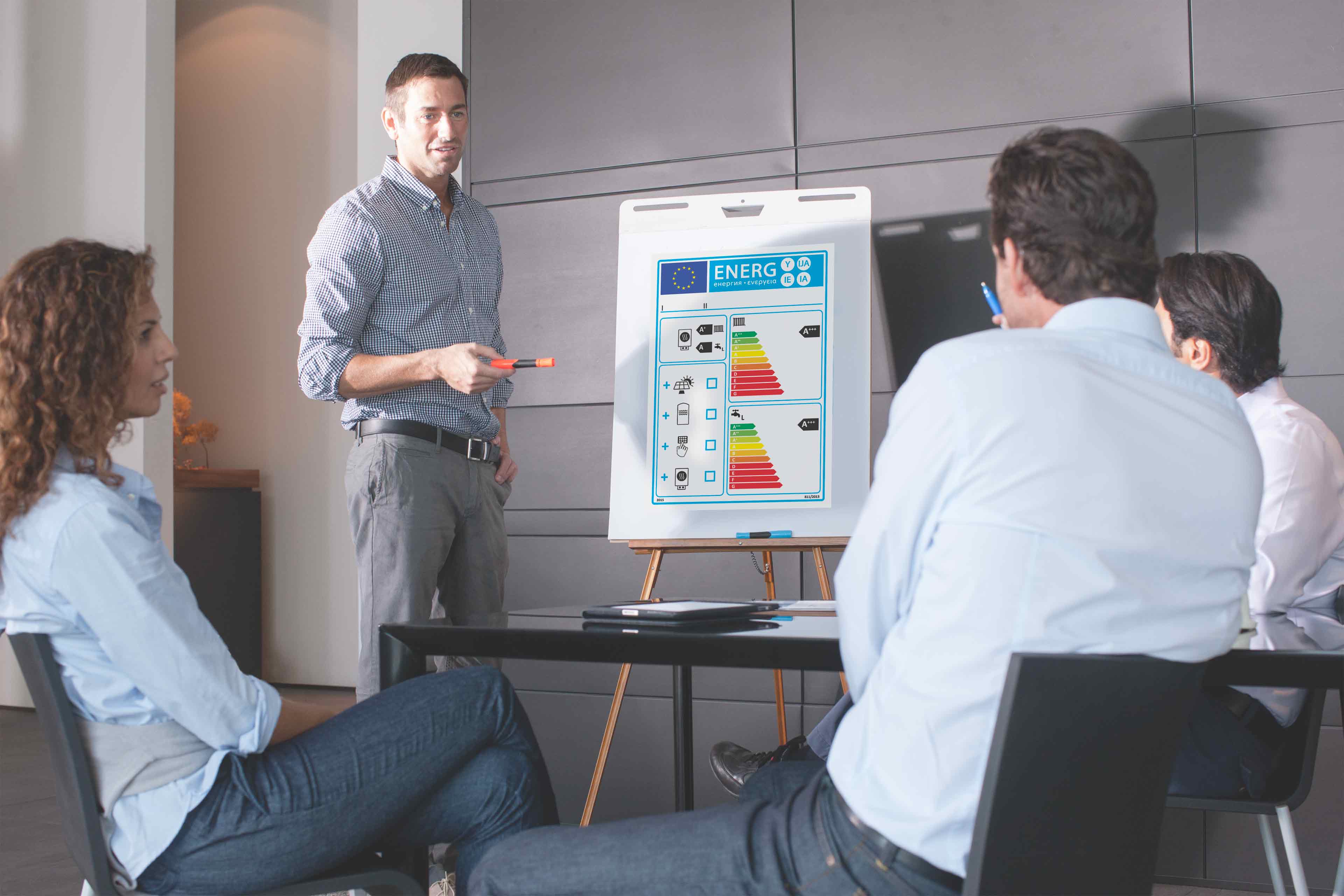 Ein Mann steht und zeigt auf eine Energieeffizienz-Tabelle auf einer Staffelei, während drei sitzende Kollegen während einer Geschäftsbesprechung in einem modernen Büro aufmerksam zuhören.