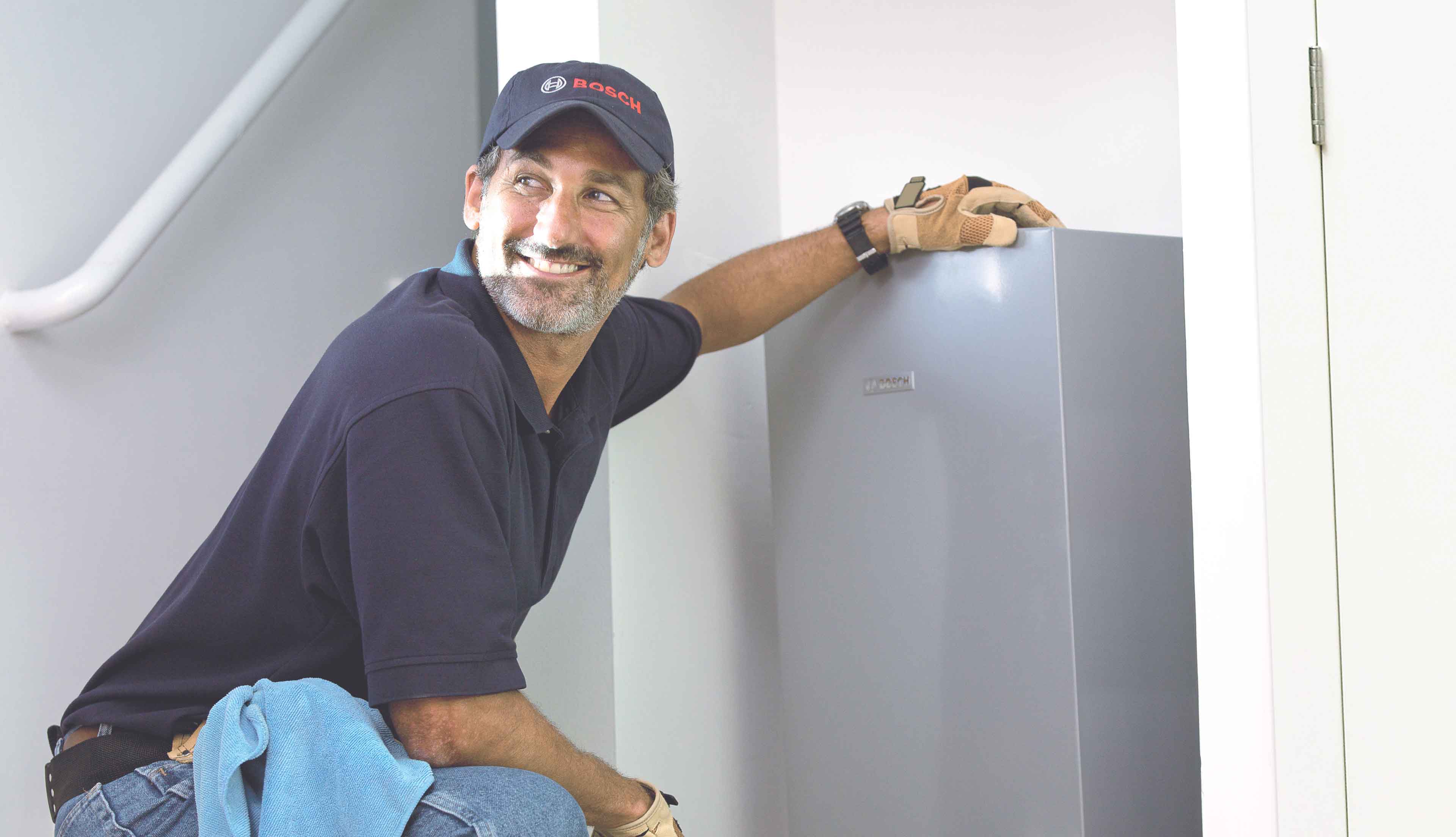 Ein lächelnder Mann mit Marinekappe und Handschuhen hockt neben einem grauen Metallschrank und scheint in einem Gebäude Wartungs- oder Installationsarbeiten für den Bosch Werkskundendienst durchzuführen.