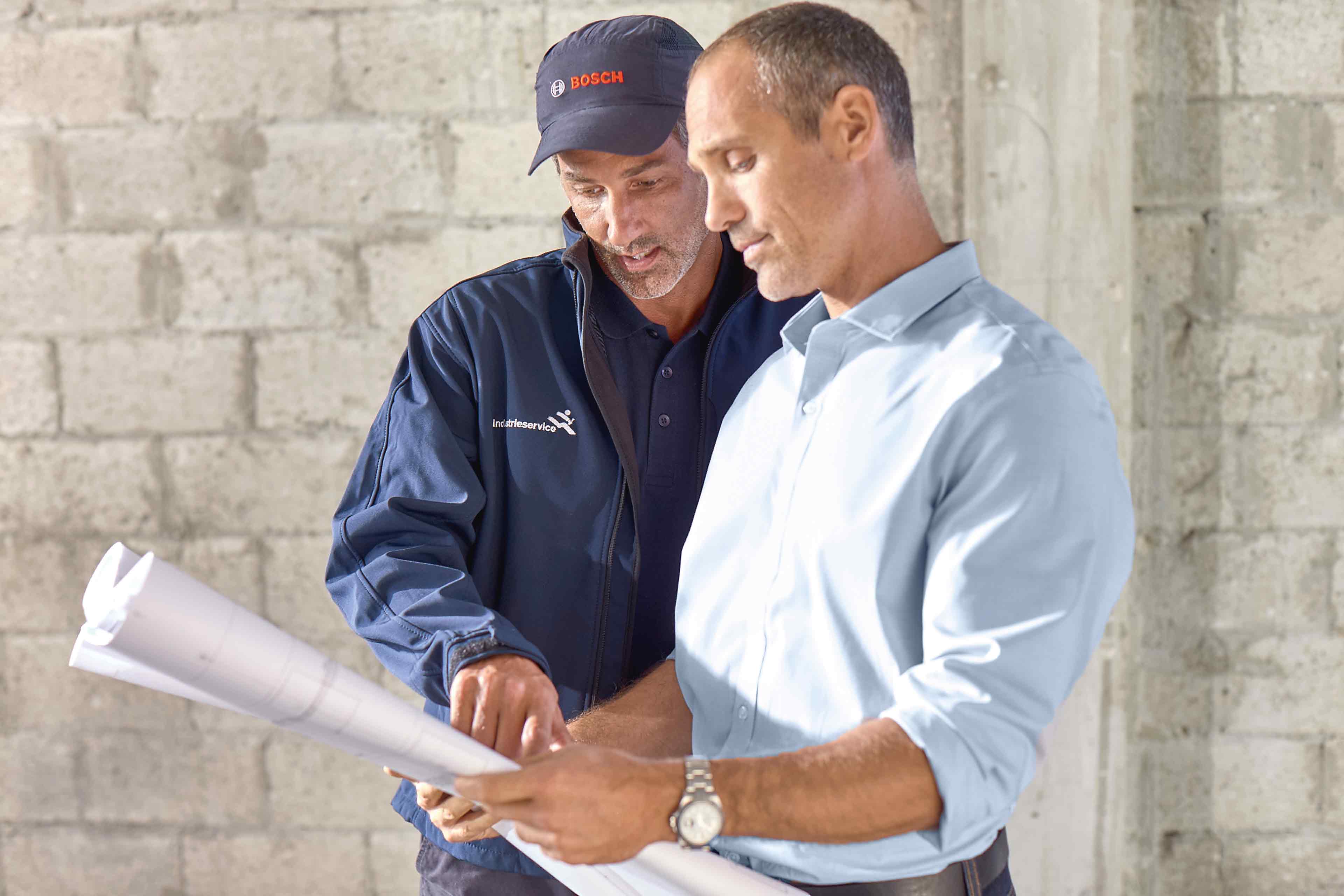 Zwei Männer stehen in einem Gebäude und betrachten Baupläne. Der eine trägt eine Bosch Werkskundendienst-Mütze und eine blaue Jacke, der andere ein hellblaues Hemd. Sie besprechen Baupläne in einem teilweise bebauten Raum.