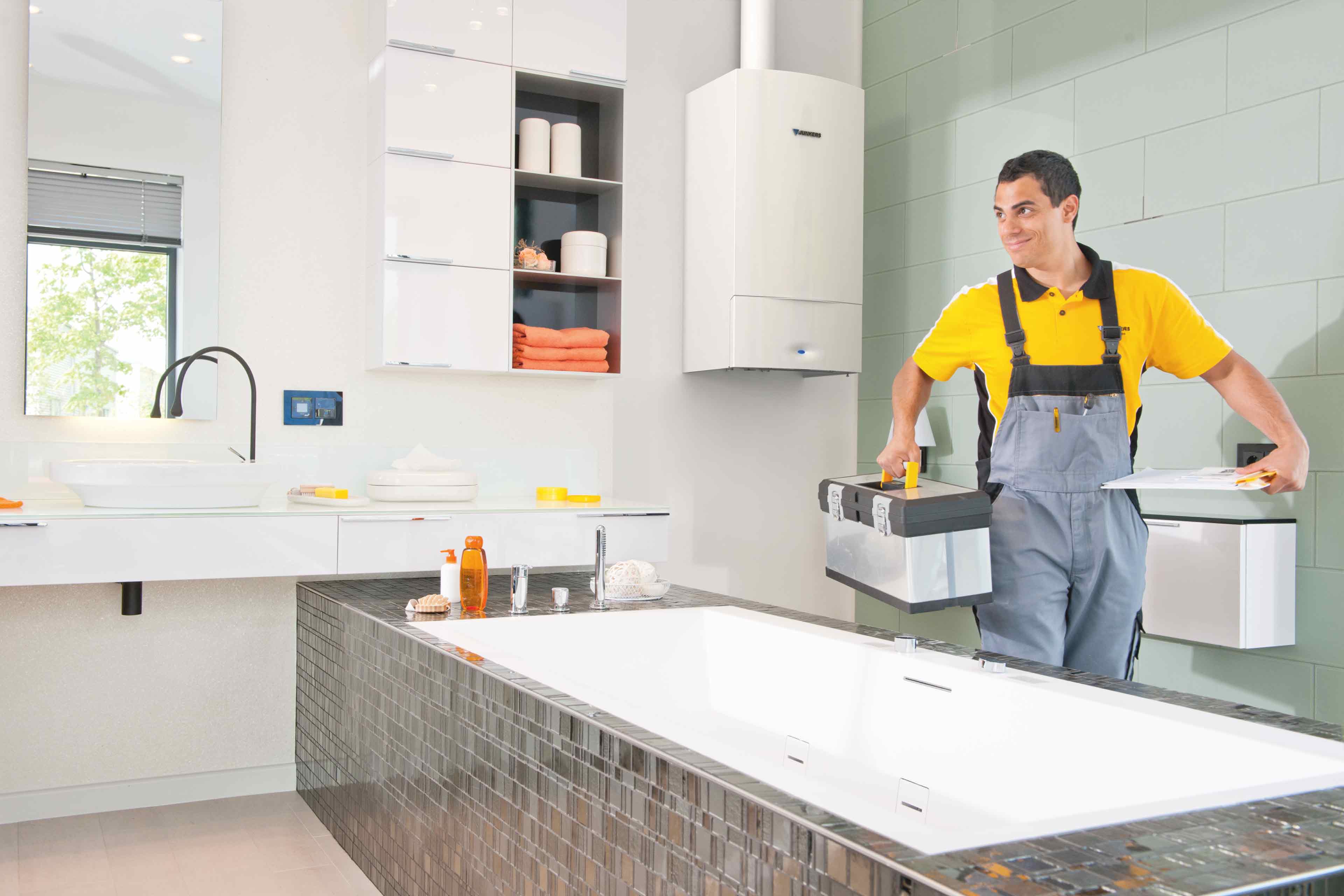 Ein Klempner in grauer Latzhose und gelbem Hemd, der den Bosch Werkskundendienst repräsentiert, trägt einen Werkzeugkasten und Geräte in ein modernes, helles Badezimmer mit einer großen Badewanne und schlichten Armaturen.