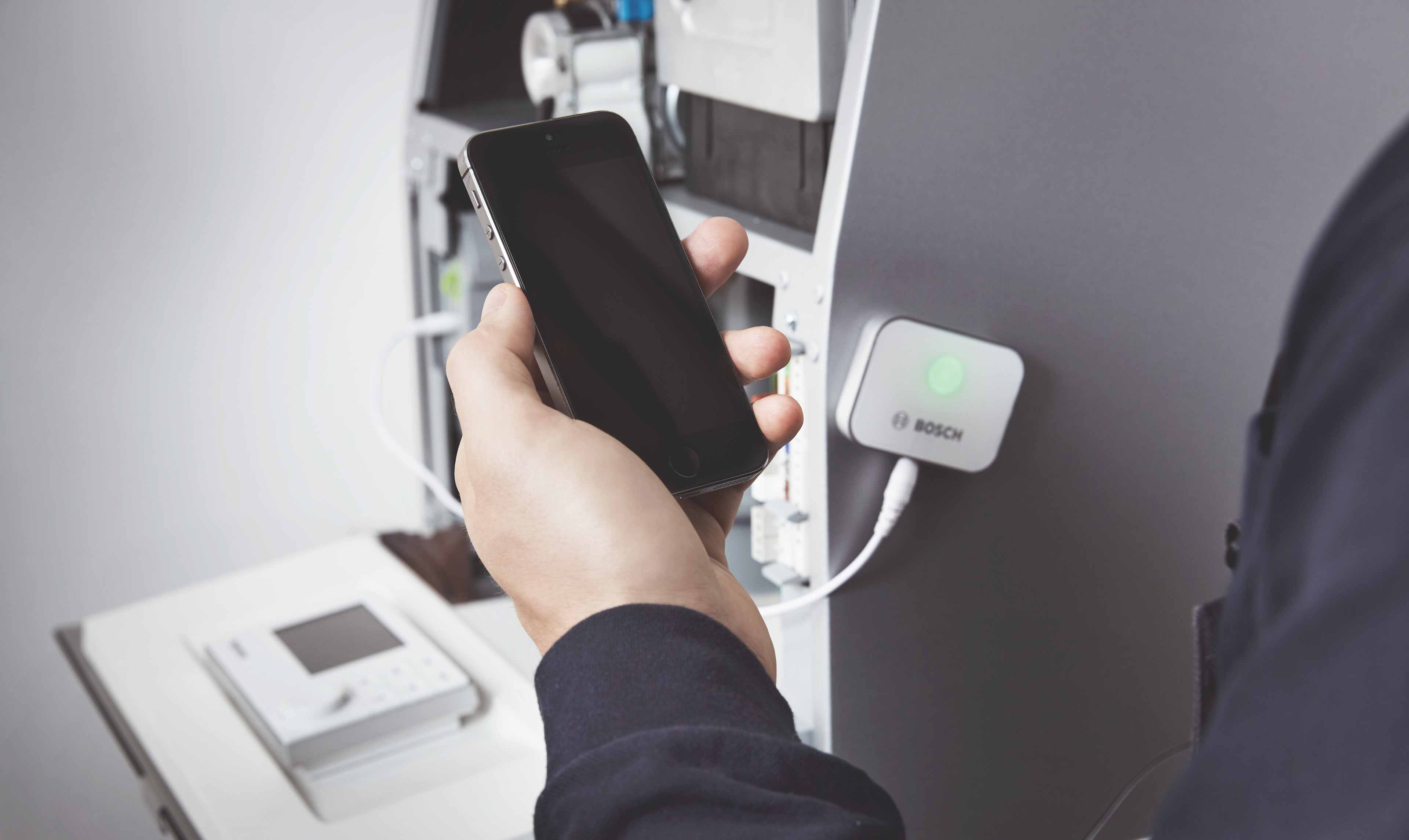 Eine Person hält ein Smartphone in die Nähe eines Bosch-Geräts mit einer grün leuchtenden Anzeige, das mit einem weißen Kabel an eine Maschine angeschlossen ist. Im Hintergrund deuten Geräte und eine Tastatur auf den Bosch Werkskundendienst hin.