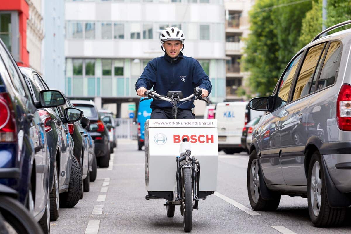 Eine Person, die einen Helm und eine dunkle Jacke tr&auml;gt, f&auml;hrt auf einem Lastenfahrrad mit der Aufschrift Bosch eine Stadtstra&szlig;e hinunter, die auf beiden Seiten von parkenden Autos ges&auml;umt ist.