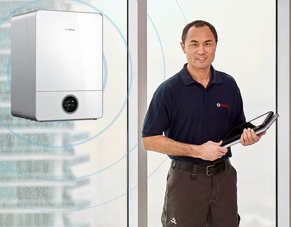 Ein Mann in Bosch-Kleidung steht in einem Haus mit einem Tablet in der Hand, neben einem Bosch-Wandgerät, mit großen Fenstern und einer Stadtlandschaft im Hintergrund.