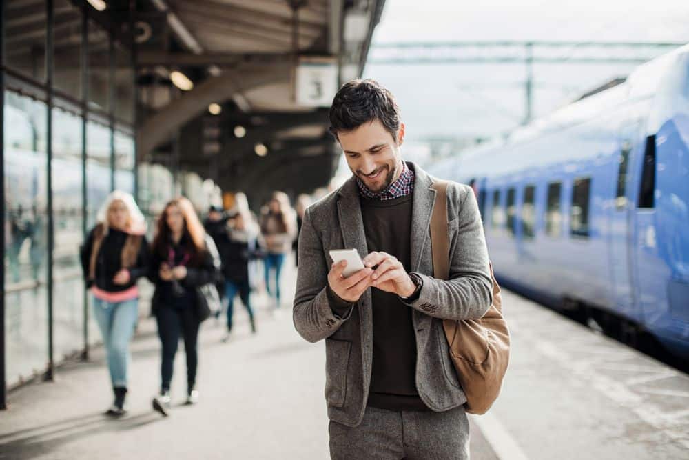 Ein Mann im Anzug steht auf einem Bahnsteig, lächelt und schaut auf sein Telefon. Er hat eine braune Tasche über der Schulter. Im Hintergrund sind ein blauer Zug und mehrere Menschen zu sehen.