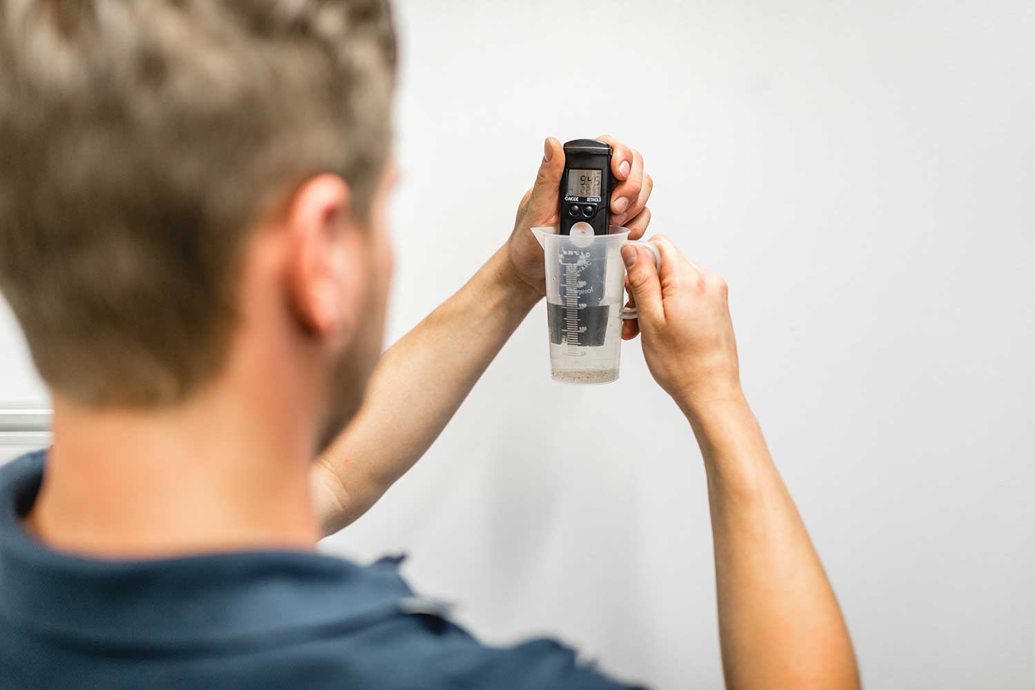 Eine Person hält ein digitales Gerät über einen durchsichtigen, mit Flüssigkeit gefüllten Messbecher und scheint vor einem schlichten weißen Hintergrund eine Messung vorzunehmen - möglicherweise im Rahmen der Qualitätskontrolle des Bosch Werkskundendienstes.