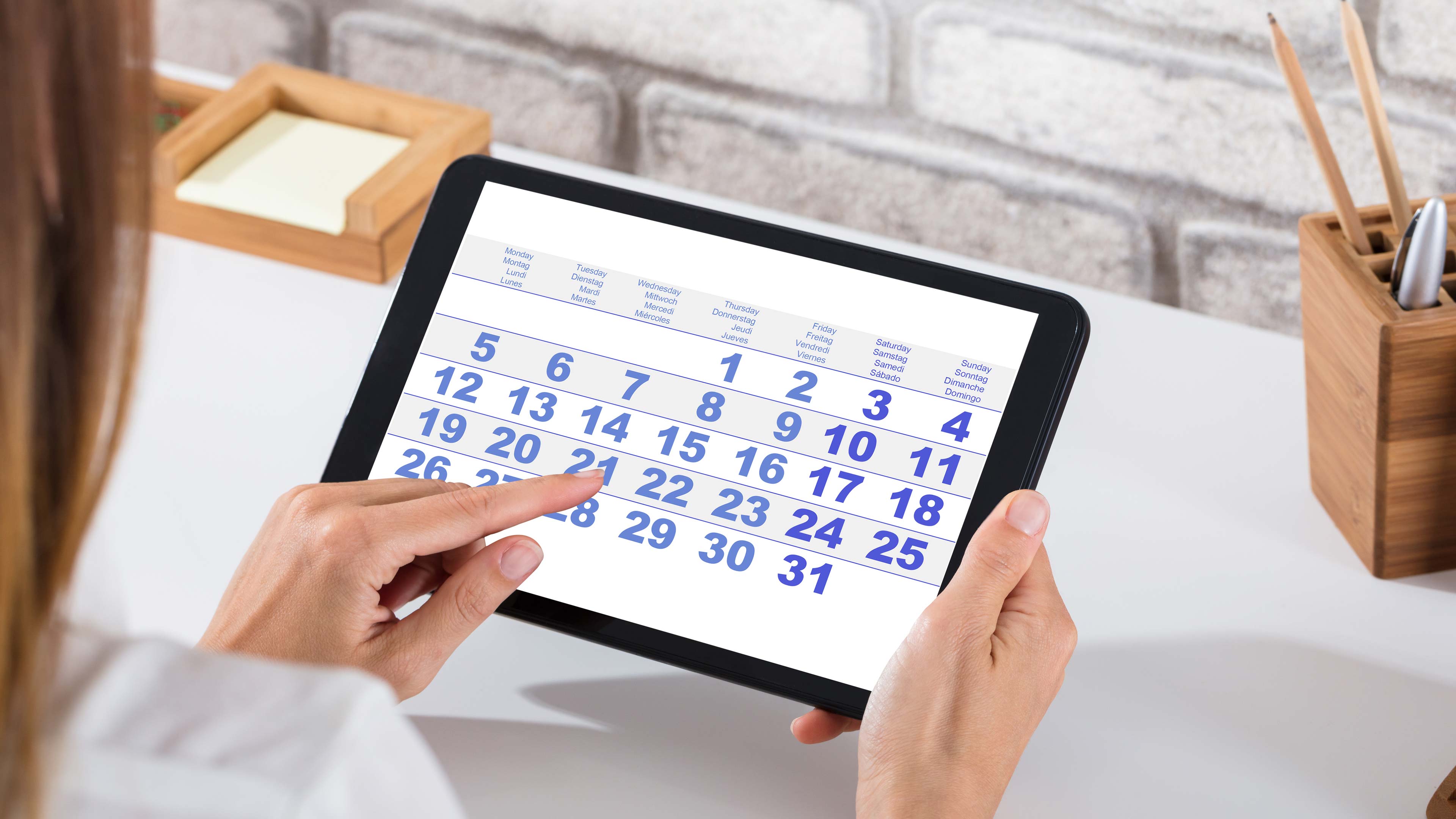 Eine Person, die ein Tablet hält, zeigt einen digitalen Kalender, wobei ihr Zeigefinger auf den 14. Der Hintergrund zeigt einen Schreibtisch mit Büromaterial und eine weiße Backsteinmauer.