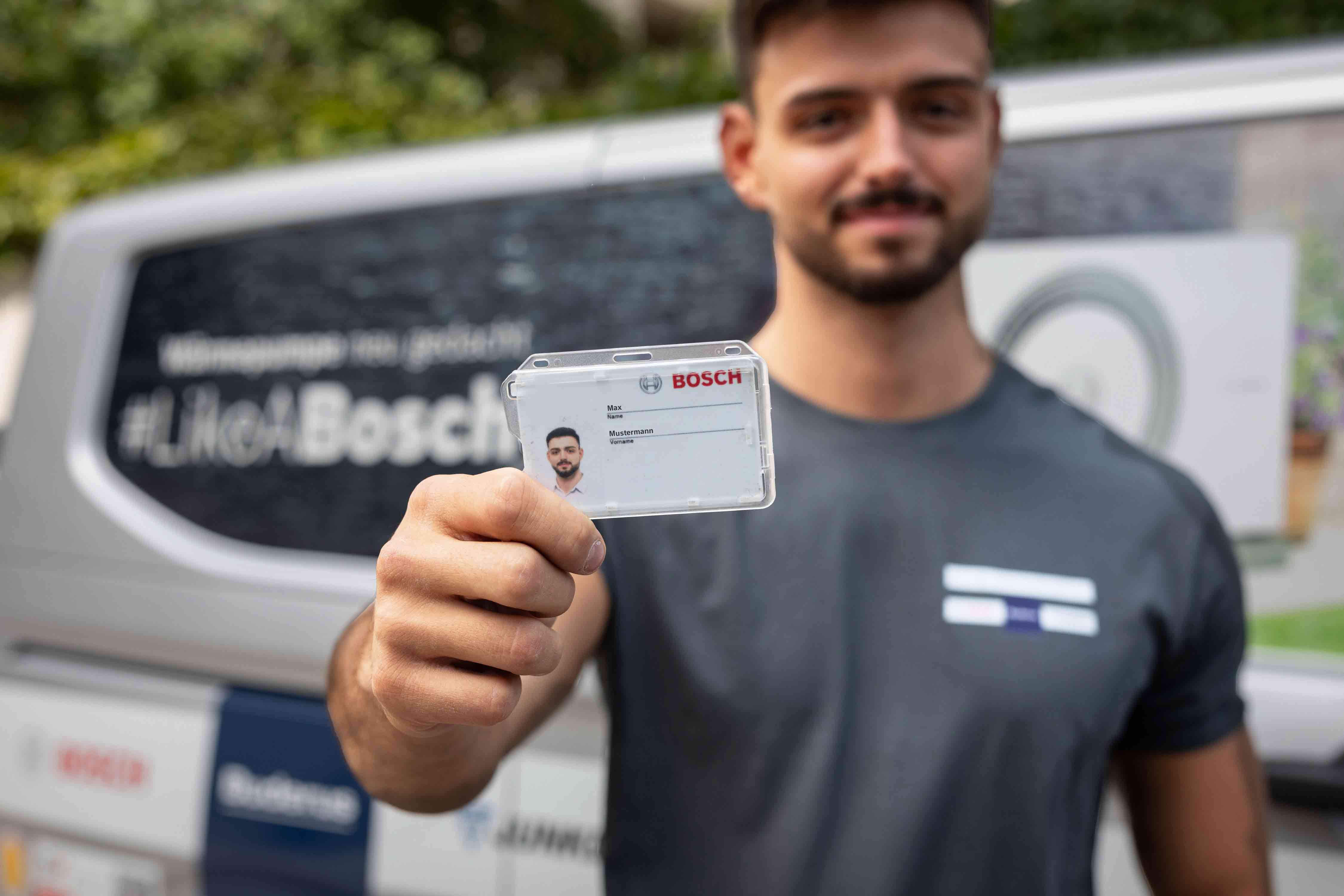 Ein Mann in einem grauen Hemd hält einen Bosch-Ausweis in die Kamera und lächelt. Im unscharfen Hintergrund ist ein Lieferwagen mit Bosch-Branding und dem Hashtag #LikeABosch zu sehen.