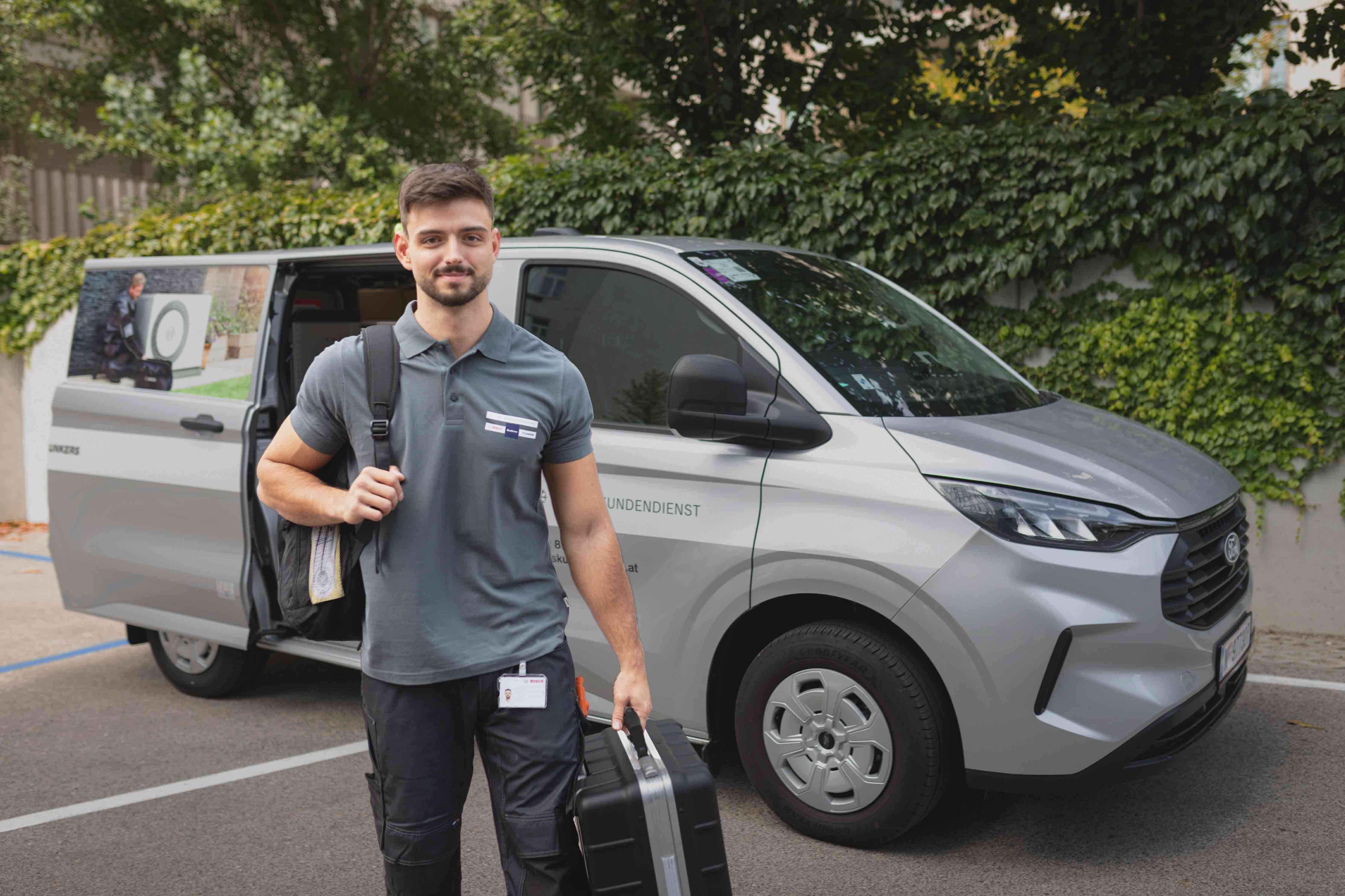 Ein Mann in grauer Uniform, der einen schwarzen Koffer und eine Tasche trägt, geht von einem grauen Thermenservice-Wagen weg, der auf der Straße geparkt ist. Der Lieferwagen hat einen Schriftzug an der Seite, im Hintergrund ist Grün zu sehen.