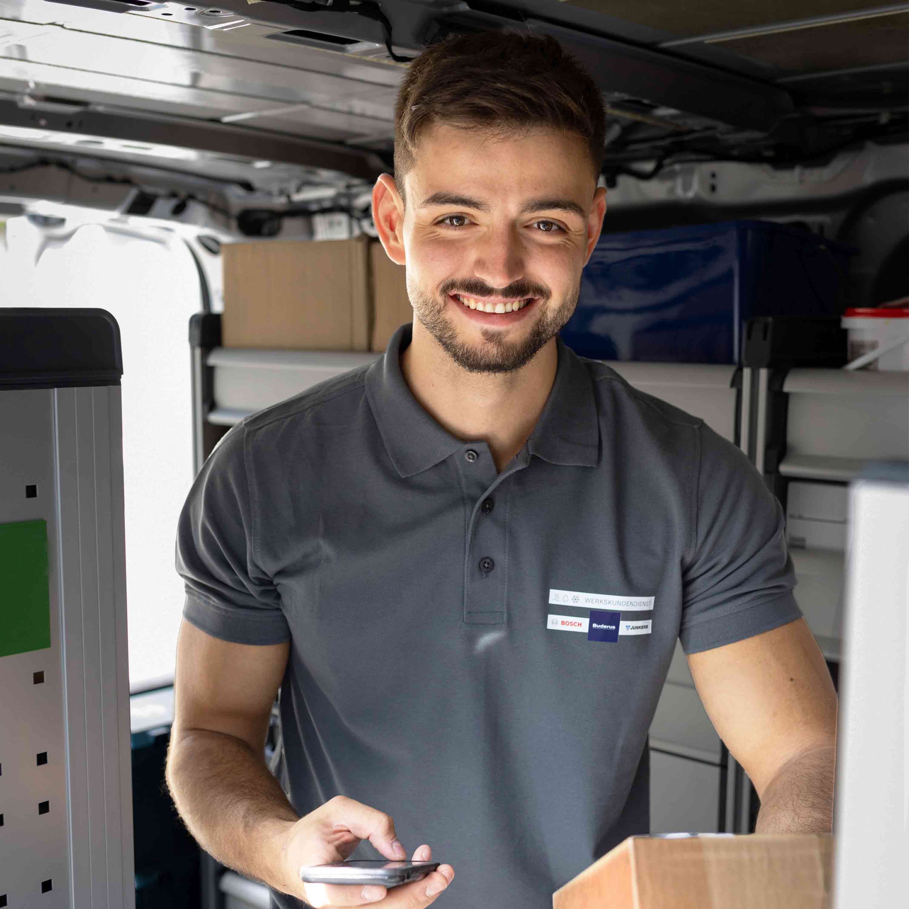 Ein lächelnder Techniker in einem grauen Poloshirt steht in einem Lieferwagen, in der Hand ein Smartphone und ein Paket, umgeben von Kisten und Regalen.