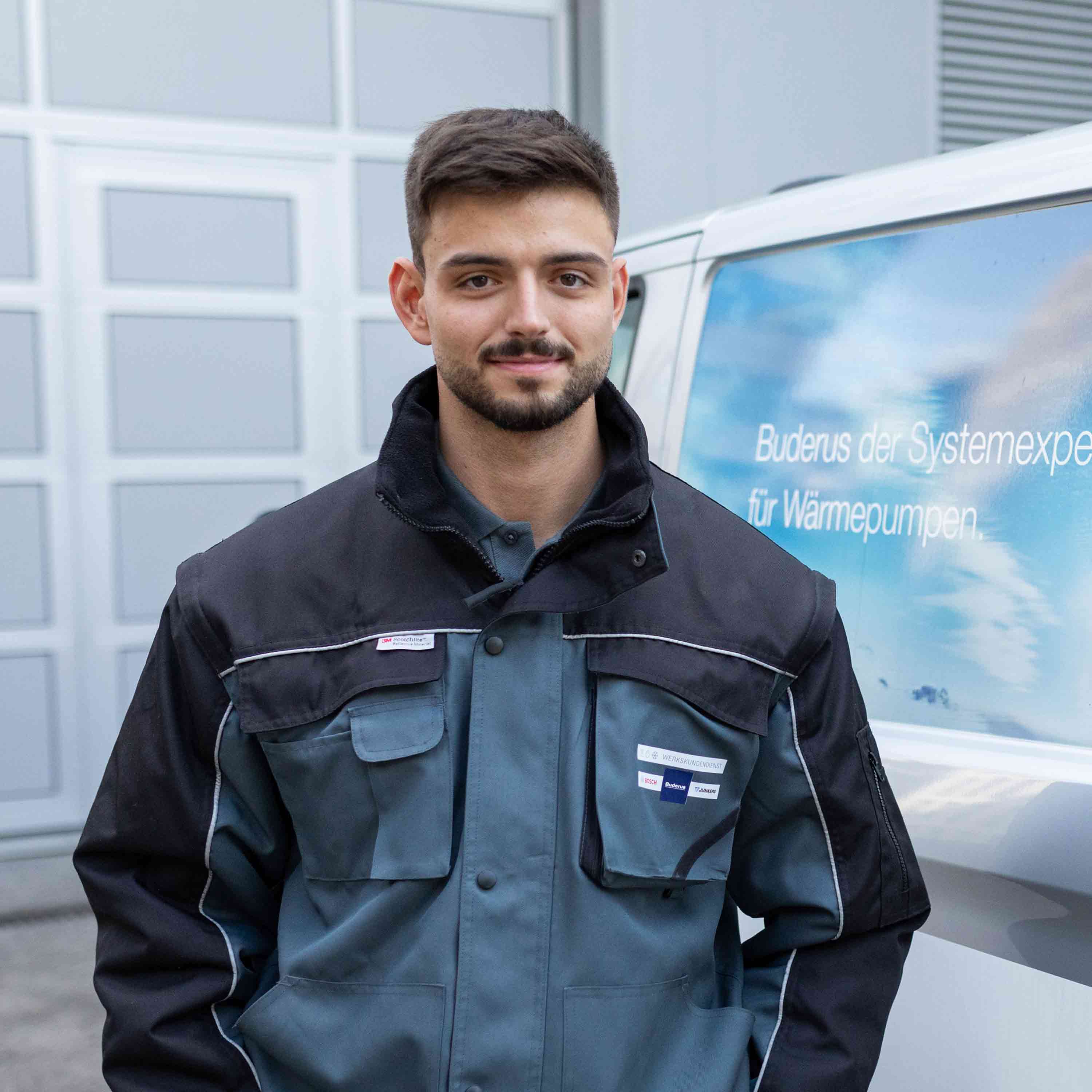 Ein junger Mann in einer blau-schwarzen Arbeitsuniform steht vor einem weißen Lieferwagen mit deutscher Aufschrift auf dem Fenster und lächelt in die Kamera.