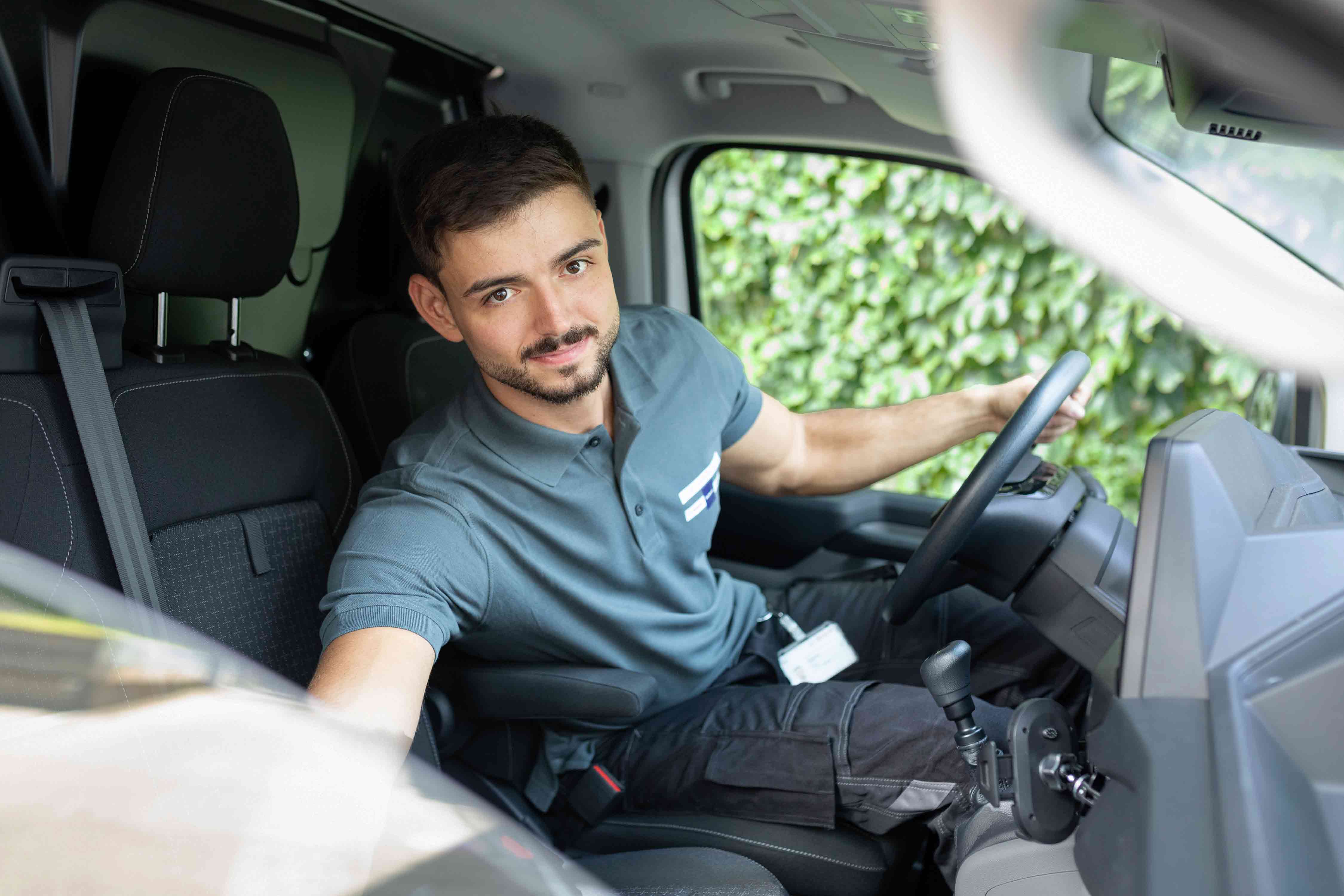 Ein Mann in einem grauen Polohemd sitzt auf dem Fahrersitz eines Fahrzeugs, blickt in die Kamera und greift zur Beifahrerseite. Durch das Fenster hinter ihm ist grünes Laub zu sehen.