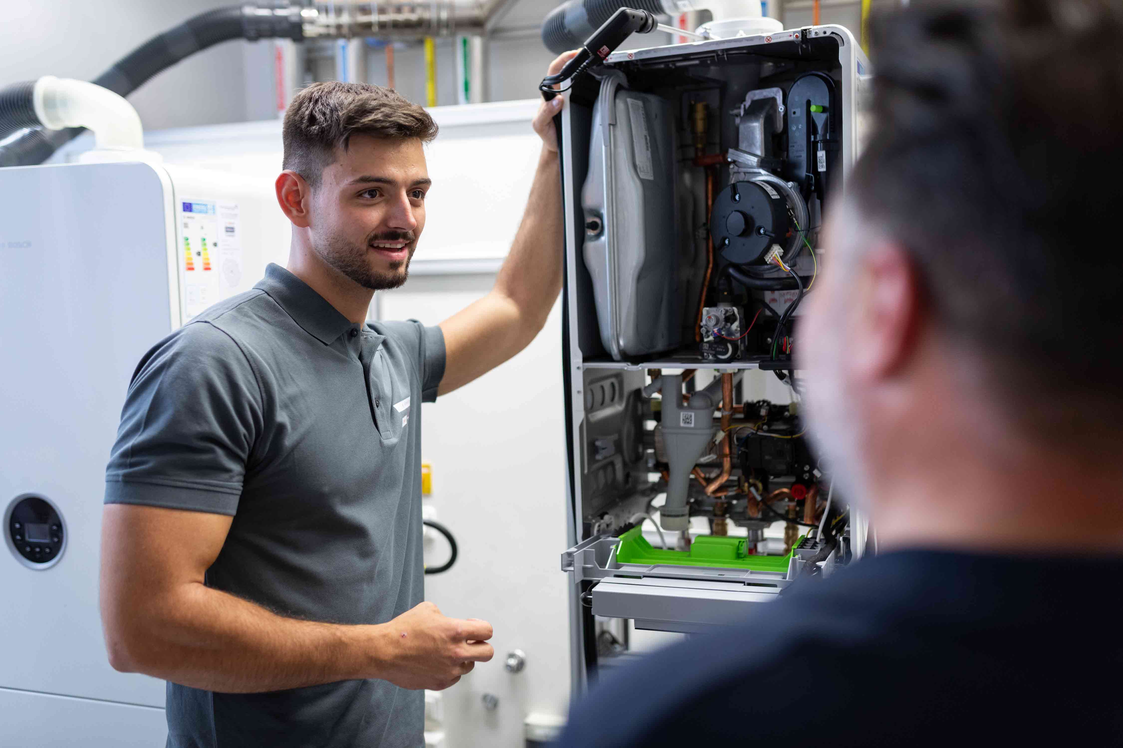 Ein Techniker erklärt die Komponenten eines offenen Kessels während einer Wartung in einer Werkstatt und deutet auf die freiliegenden Maschinen im Inneren des Geräts.