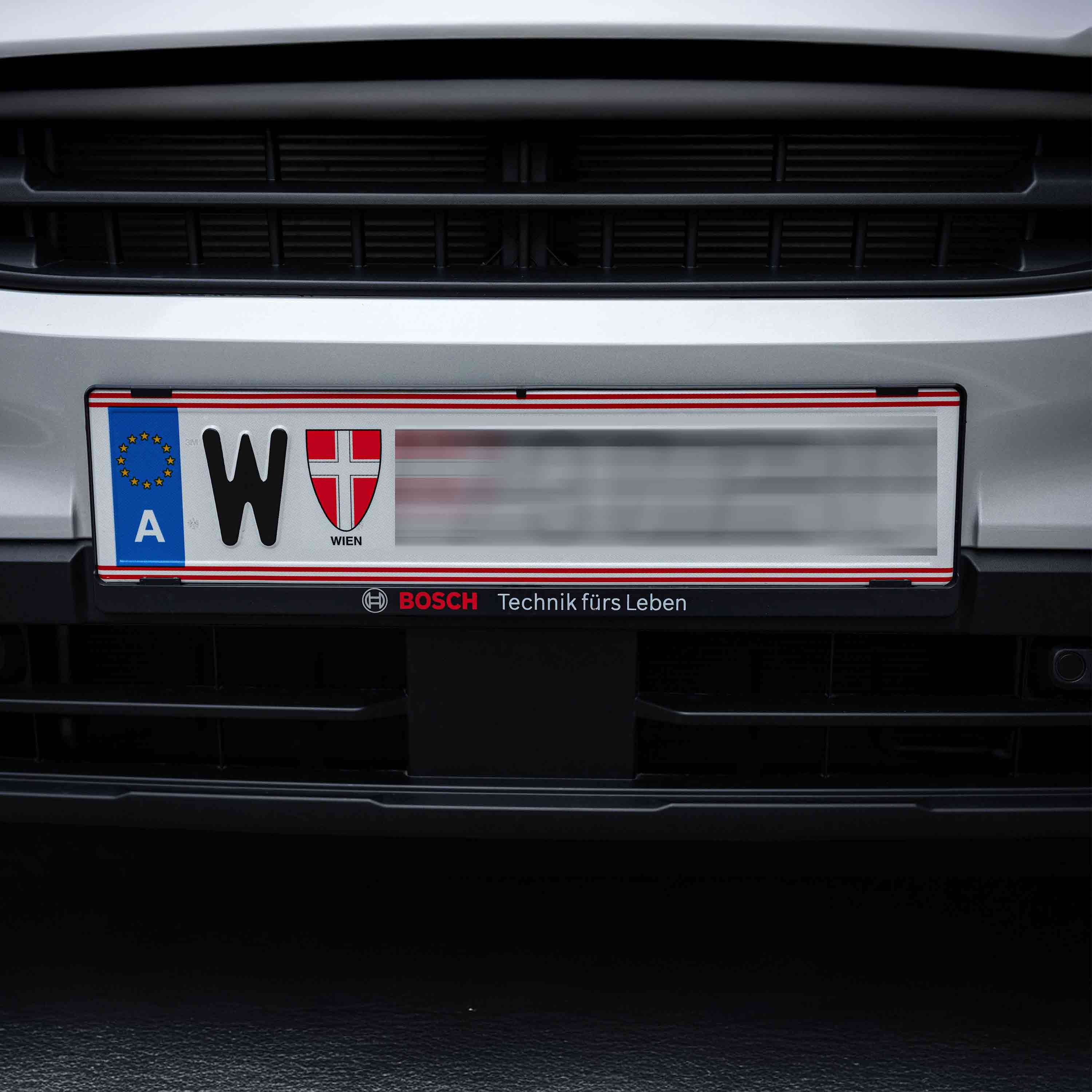 Eine Nahaufnahme eines europäischen Kfz-Kennzeichens aus Wien, Österreich, das an der Vorderseite eines silbernen Fahrzeugs angebracht ist. Das Schild zeigt ein EU-Symbol, den Buchstaben W und ein rot-weißes Wappen. Die Zahlen sind unscharf.