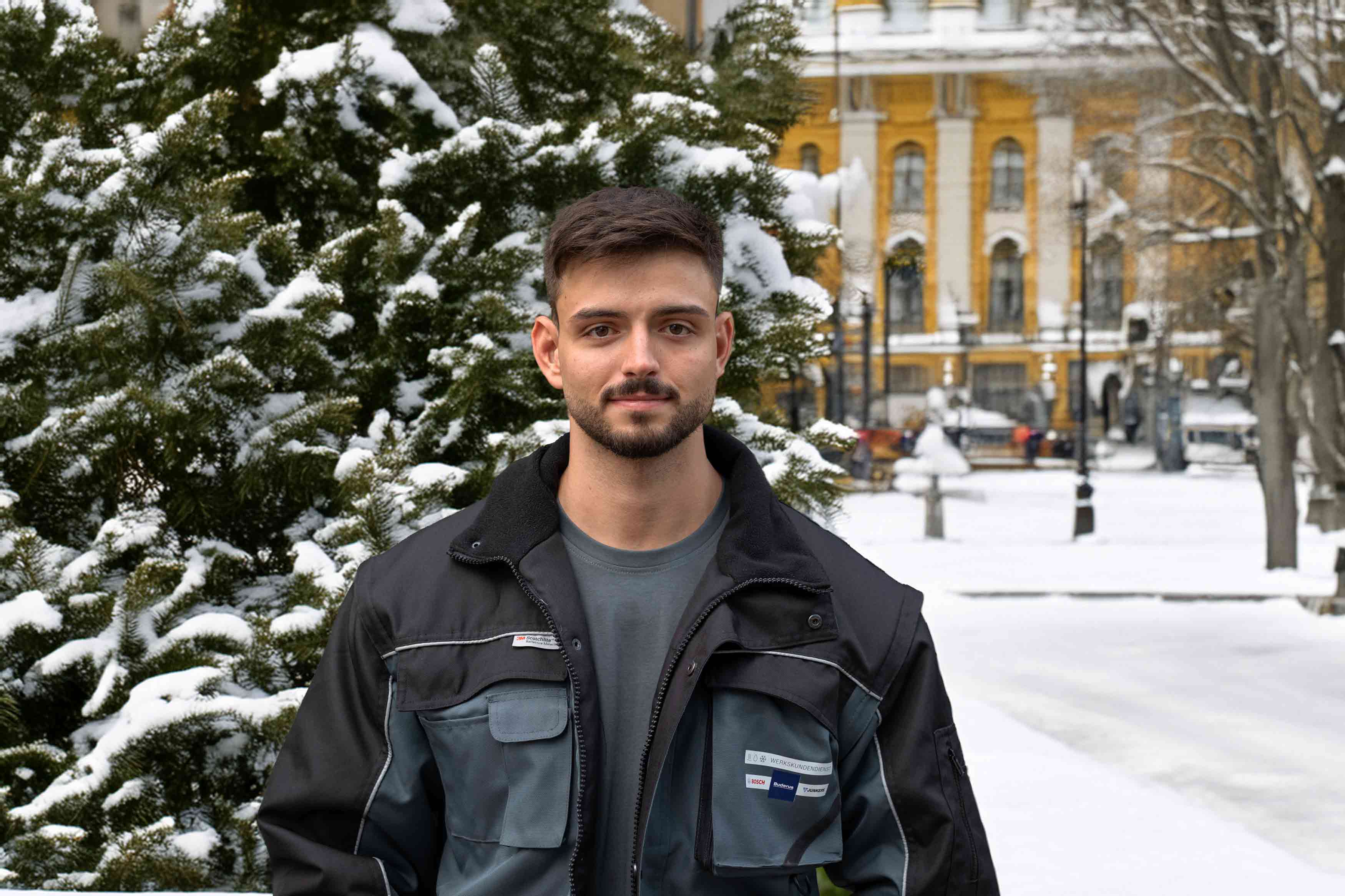Ein junger Mann mit kurzen dunklen Haaren und Bart steht im Freien vor einem schneebedeckten, immergrünen Baum und trägt eine schwarz-graue Bosch Werkskundendienst-Jacke. Im Hintergrund sind ein gelbes Gebäude und ein verschneiter Park zu sehen.