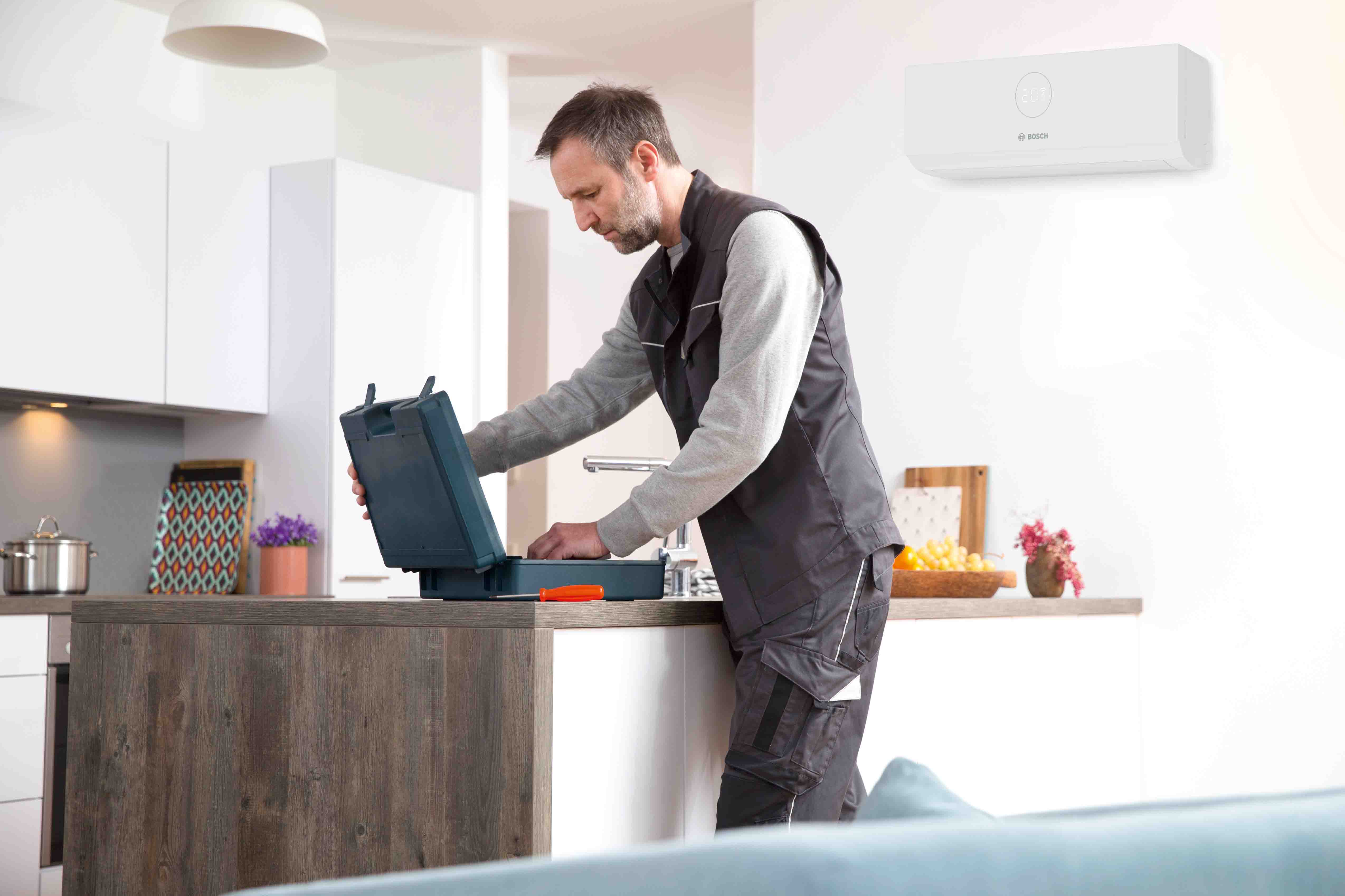 Ein Mann in Arbeitskleidung steht an einer K&uuml;cheninsel und &ouml;ffnet einen schwarzen Werkzeugkasten. Hinter ihm ist in einer hellen und aufger&auml;umten K&uuml;che ein modernes wei&szlig;es Klimager&auml;t an der Wand montiert.