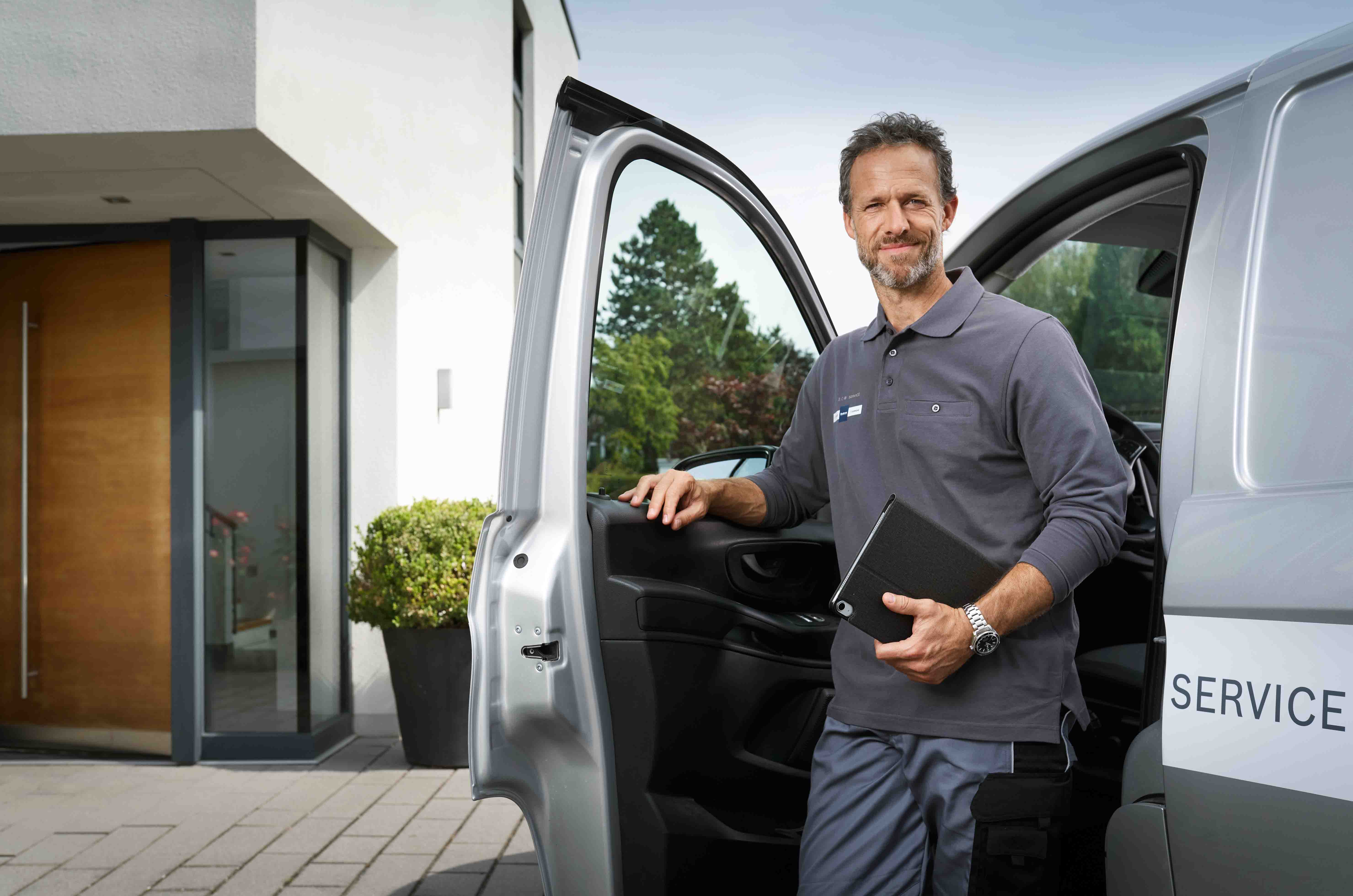 Ein l&auml;chelnder Mann in einer Dienstuniform steht an der offenen T&uuml;r eines Lieferwagens und h&auml;lt einen Ordner in der Hand. Er scheint bereit f&uuml;r die Arbeit zu sein. Im Hintergrund sind ein moderner Geb&auml;udeeingang und Gr&uuml;nfl&auml;chen zu sehen.
