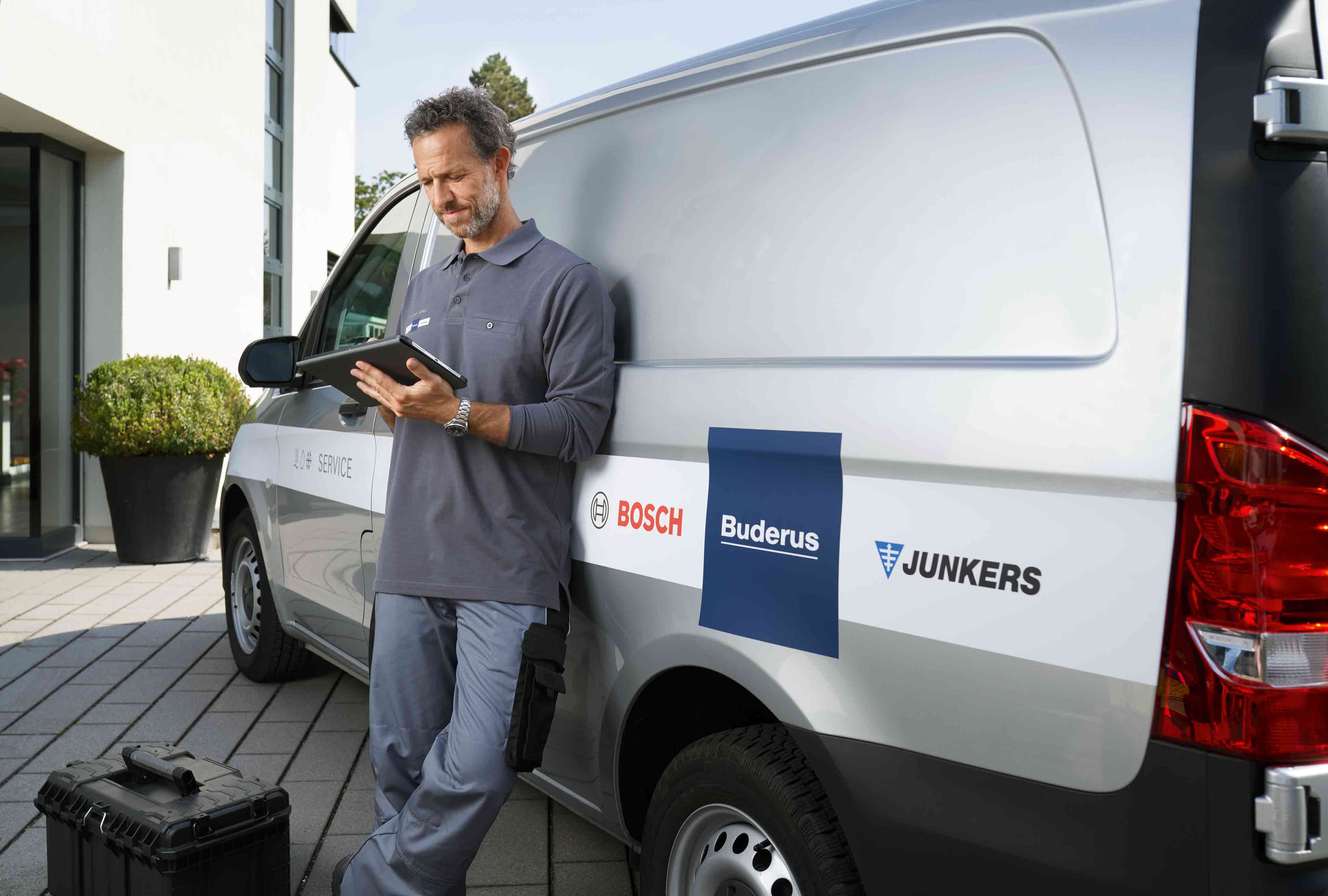 Ein Techniker in einer grauen Uniform steht an einem Servicewagen und benutzt ein Tablet. Der Wagen tr&auml;gt die Logos von Bosch, Buderus und Junkers. Ein schwarzer Werkzeugkoffer liegt in der N&auml;he auf dem Boden.