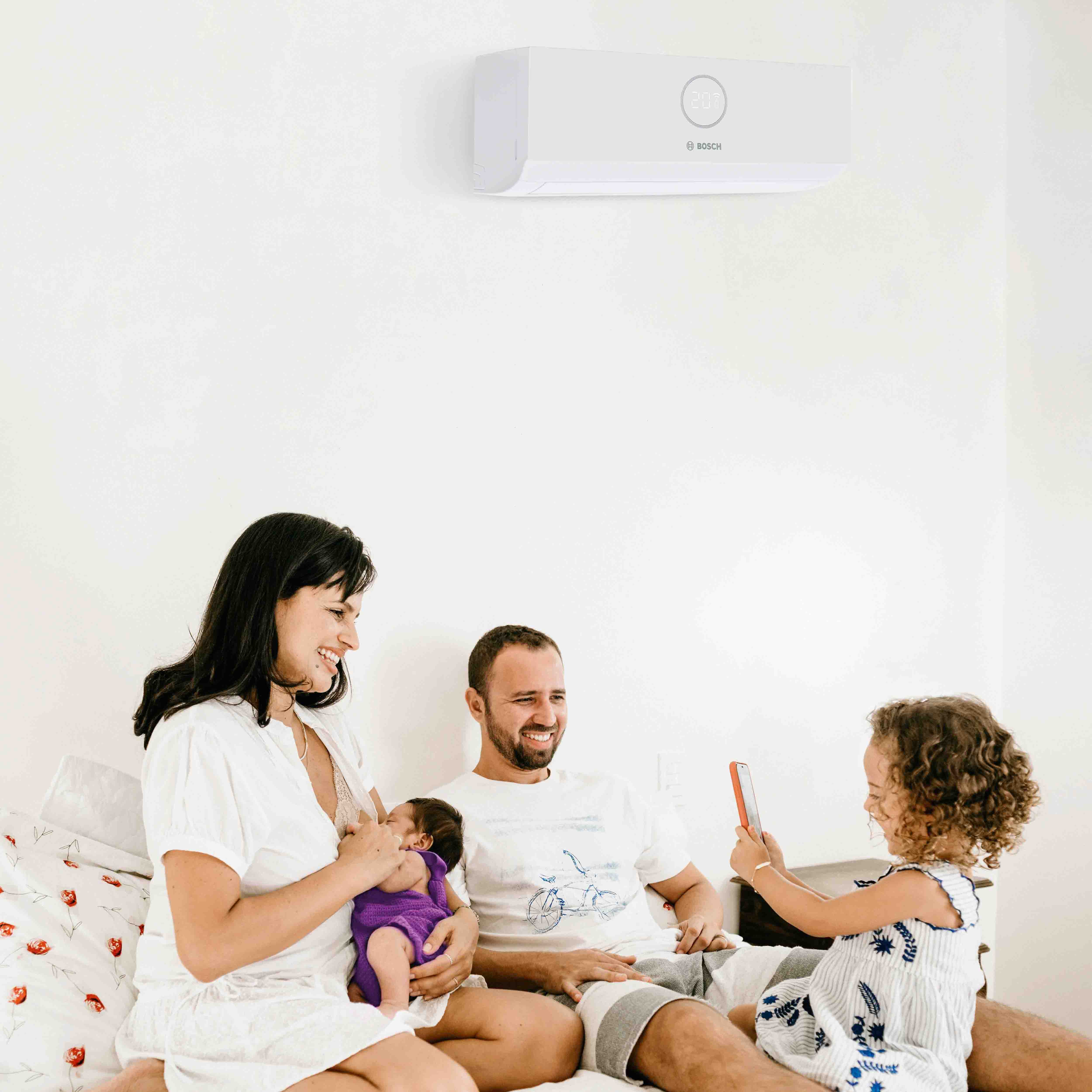 Eine vierk&ouml;pfige Familie sitzt auf einem Bett unter einer wandmontierten Klimaanlage. Die Mutter h&auml;lt ein Baby im Arm, der Vater l&auml;chelt, und ein junges M&auml;dchen macht ein Foto mit einem Telefon. Alle wirken gl&uuml;cklich und entspannt.