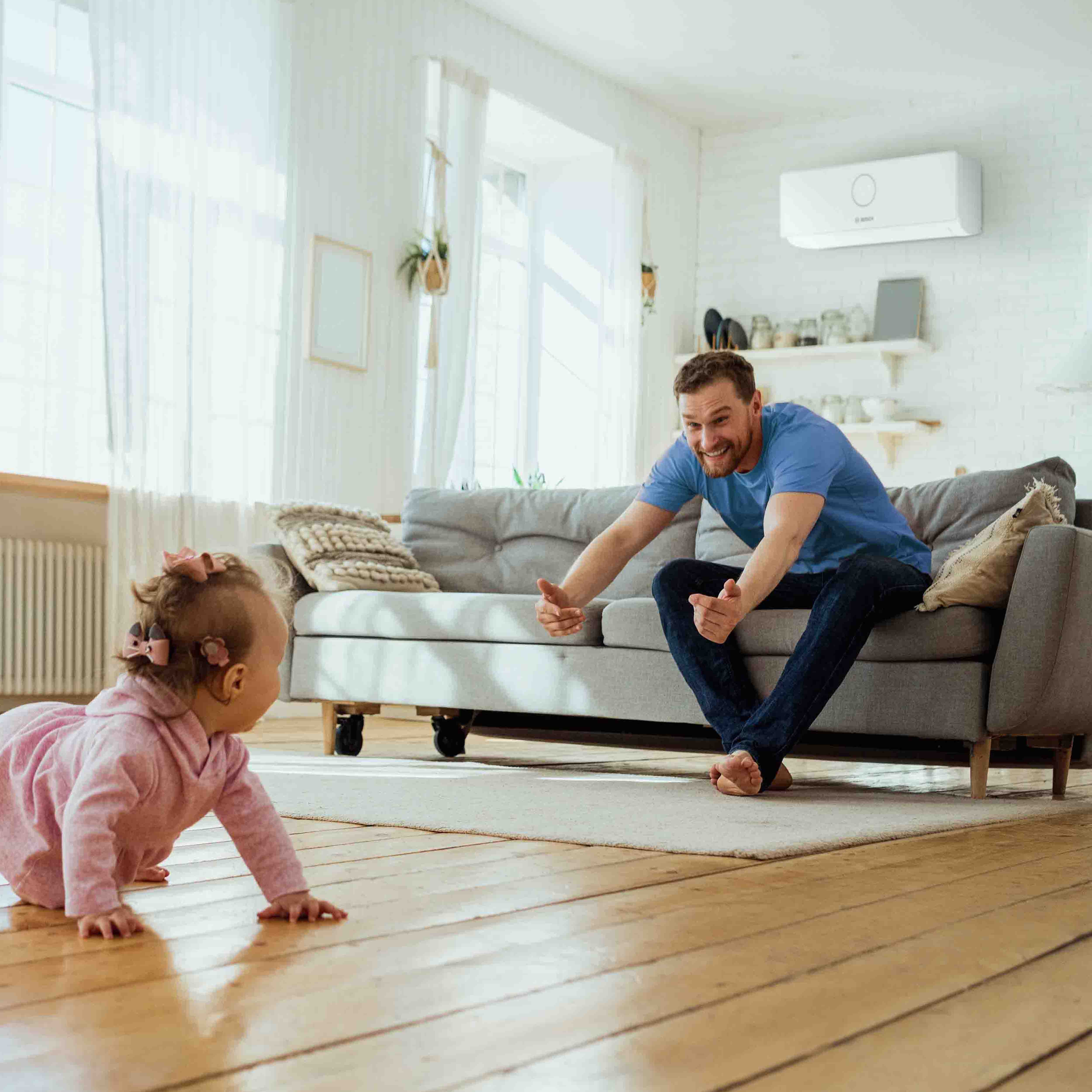 Ein Baby in rosafarbenen Kleidern krabbelt auf dem Holzboden in Richtung eines l&auml;chelnden Mannes, der auf einem grauen Sofa sitzt und es mit offenen Armen in einem hellen, gem&uuml;tlichen Wohnzimmer mit gro&szlig;en Fenstern und einer Klimaanlage an der Wand aufmuntert.