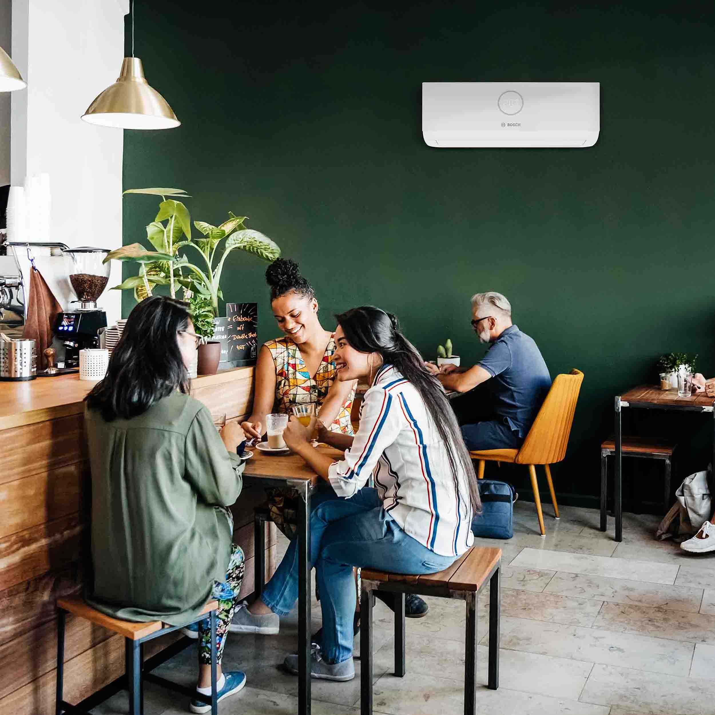 Drei Frauen sitzen an einem Holztresen in einem gem&uuml;tlichen Caf&eacute; mit gr&uuml;nen W&auml;nden, Pflanzen und einer Klimaanlage dar&uuml;ber und unterhalten sich. Ein Mann sitzt allein an einem Tisch im Hintergrund und schaut auf sein Handy.