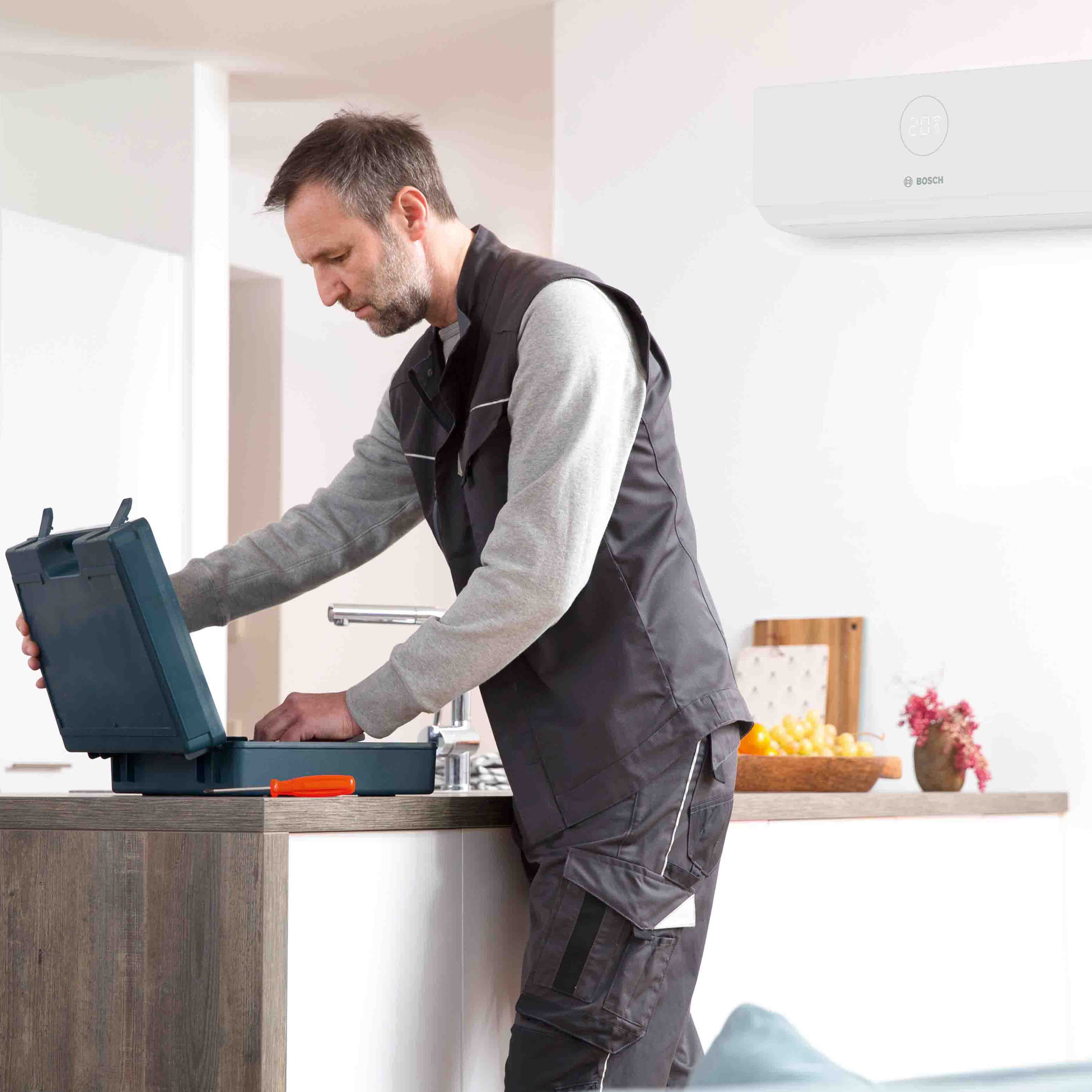 Ein m&auml;nnlicher Techniker in Arbeitskleidung steht an einem K&uuml;chentisch und &ouml;ffnet einen schwarzen Werkzeugkasten. An der Wand im Hintergrund ist ein Klimager&auml;t montiert.
