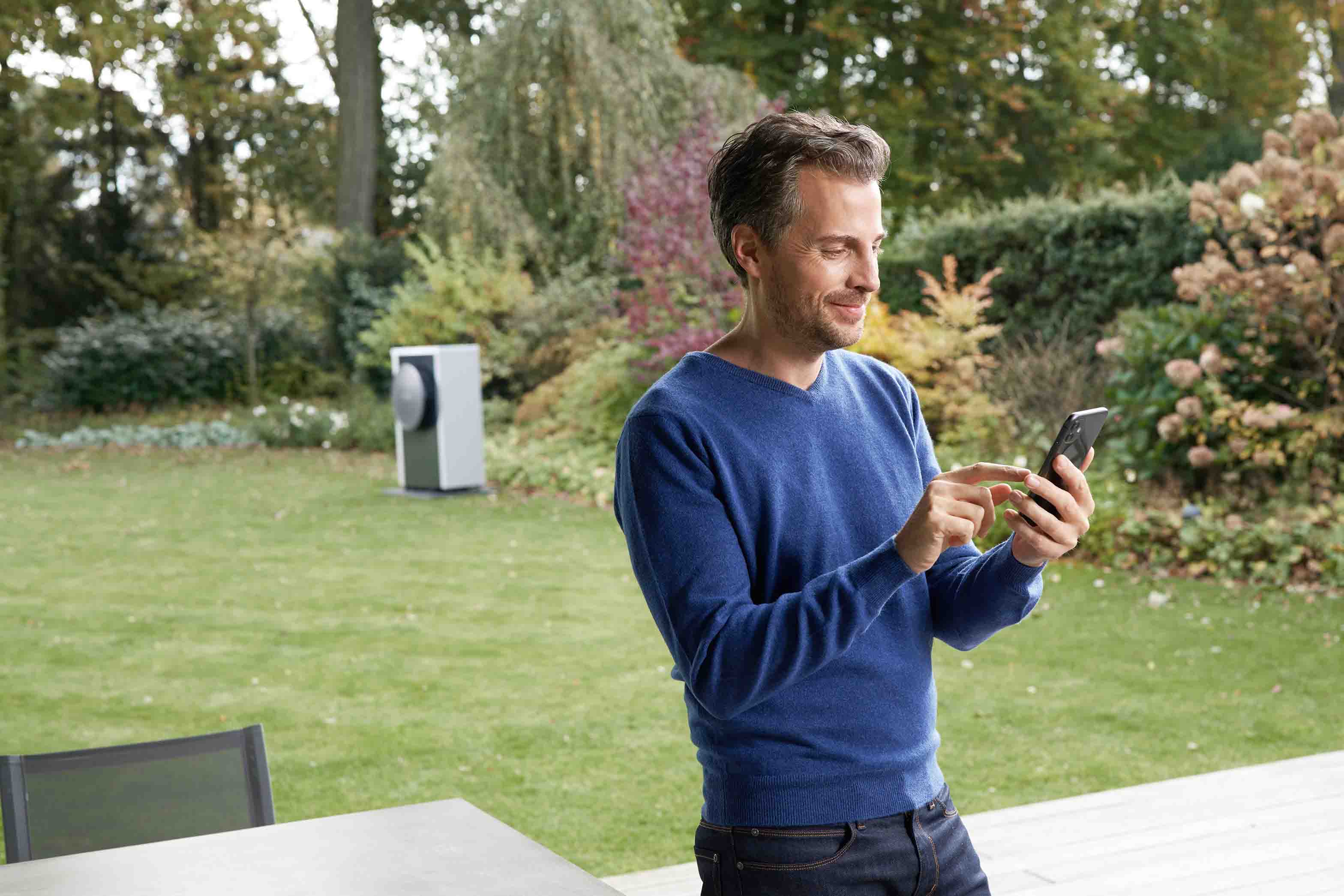 Ein Mann in einem blauen Pullover steht auf einer Terrasse, l&auml;chelt und benutzt sein Smartphone. Hinter ihm liegt ein &uuml;ppiger gr&uuml;ner Garten mit B&auml;umen und Str&auml;uchern. Im Hintergrund ist ein modernes Outdoor-Ger&auml;t zu sehen.