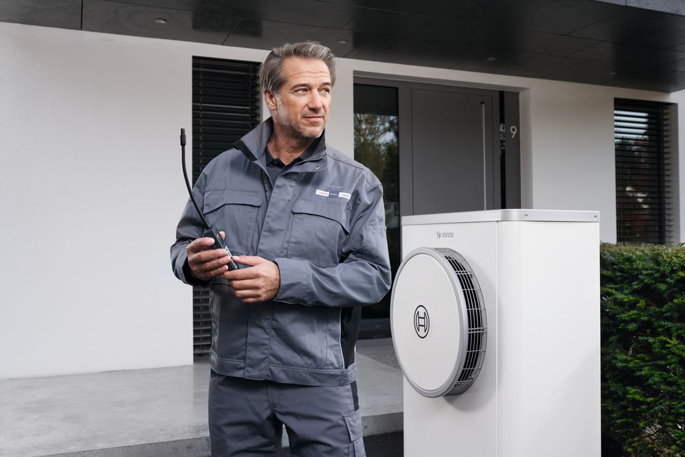 Ein Mann in grauer Uniform steht vor einem modernen Geb&auml;ude mit Werkzeugen in der Hand neben einem wei&szlig;en Bosch W&auml;rmepumpenger&auml;t.