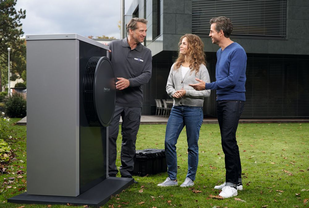 Ein Bosch Werkskundendienst-Techniker erkl&auml;rt einem l&auml;chelnden Mann und einer l&auml;chelnden Frau, die auf einer Wiese vor einem modernen Geb&auml;ude stehen, ein W&auml;rmepumpen-Au&szlig;enger&auml;t.