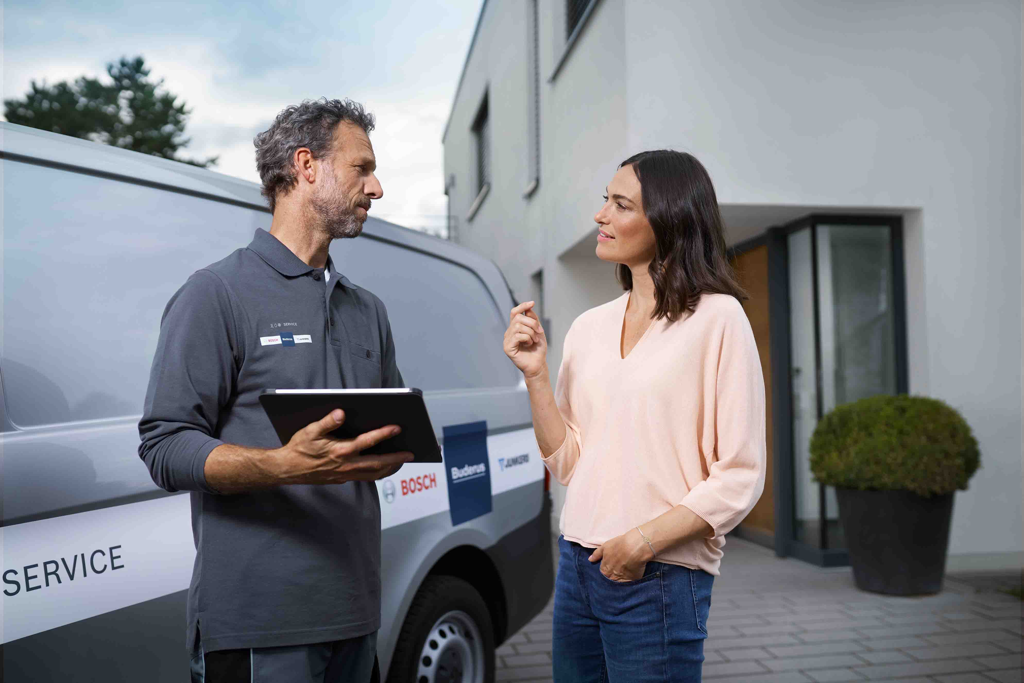 Ein Techniker, der ein Tablet in der Hand h&auml;lt, spricht mit einer Frau vor einem modernen Geb&auml;ude, das neben einem Servicewagen steht. Die Frau scheint dem Techniker eine Frage zu stellen oder etwas mit ihm zu besprechen.
