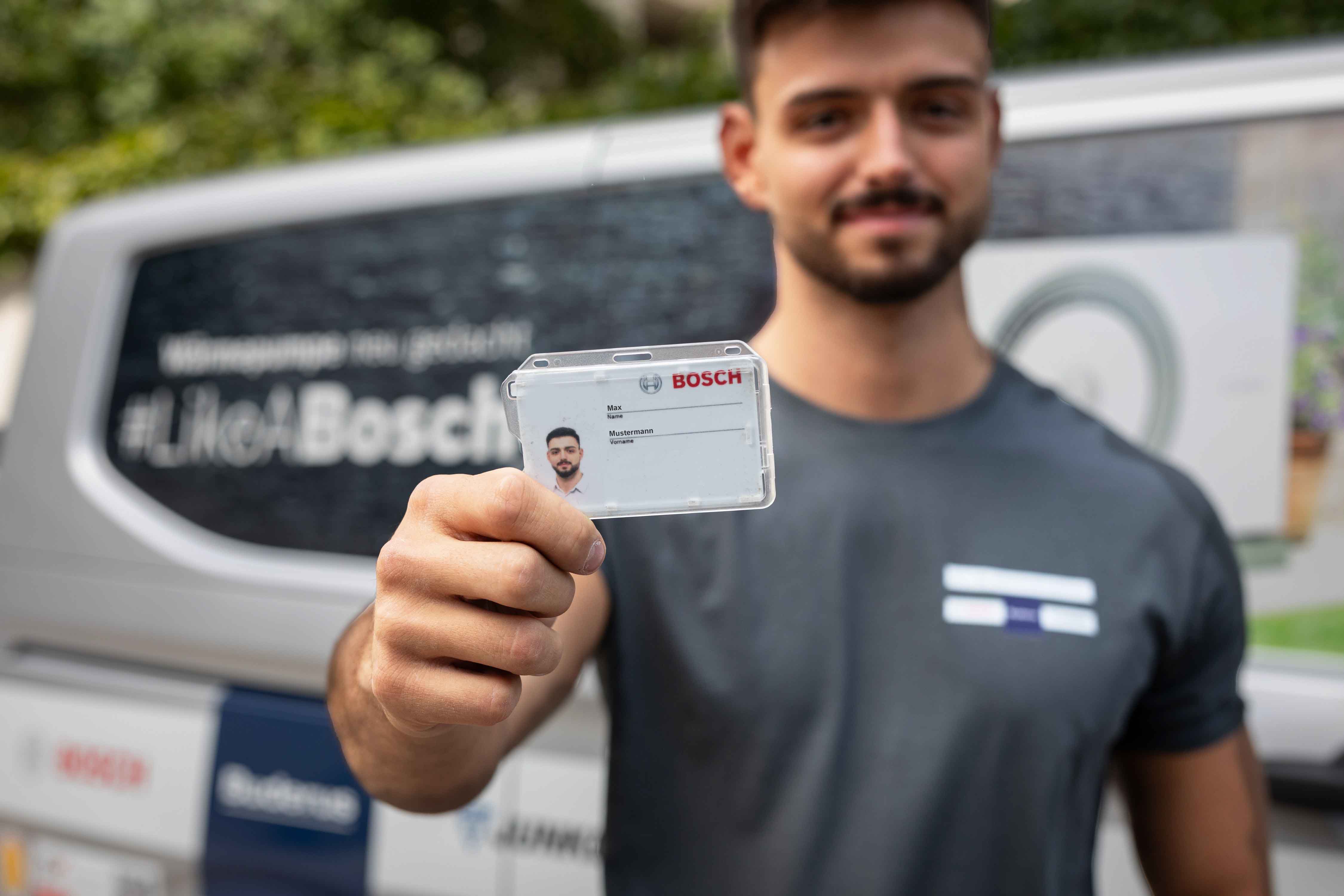 Ein Mann in einem grauen Hemd h&auml;lt einen Bosch-Mitarbeiterausweis in die Kamera. Er steht vor einem Lieferwagen mit Bosch-Logo und einem Schriftzug, der auf die innovative W&auml;rmepumpen-Technologie im Hintergrund hinweist.