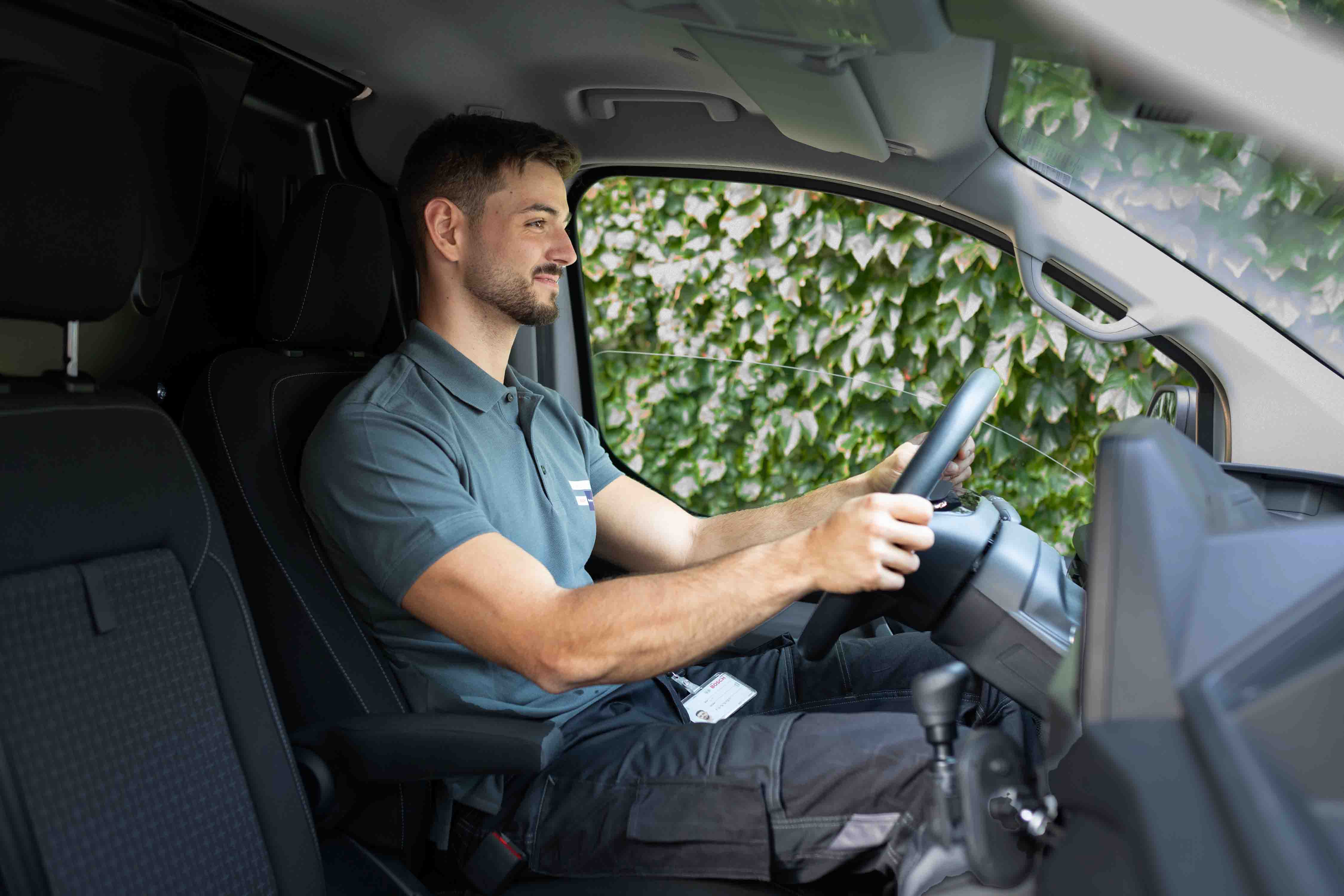 Ein Mann in einem grauen Polohemd und einer schwarzen Hose sitzt auf dem Fahrersitz eines Fahrzeugs, l&auml;chelt und h&auml;lt das Lenkrad fest. Durch das Fenster drau&szlig;en sind gr&uuml;ne Blattpflanzen zu sehen.
