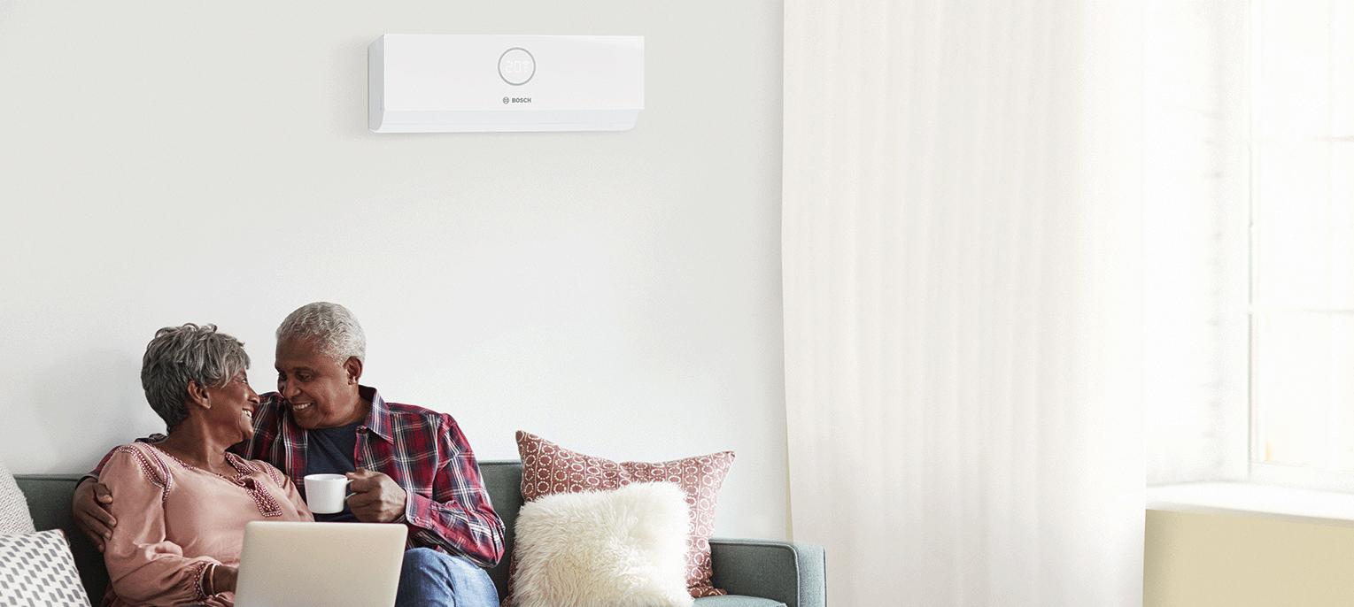 Ein &auml;lteres Ehepaar sitzt l&auml;chelnd mit einer Tasse in der Hand auf einem Sofa; &uuml;ber ihnen ist eine moderne Klimaanlage an der Wand angebracht. Durch die transparenten Vorh&auml;nge eines nahegelegenen Fensters f&auml;llt Licht herein.