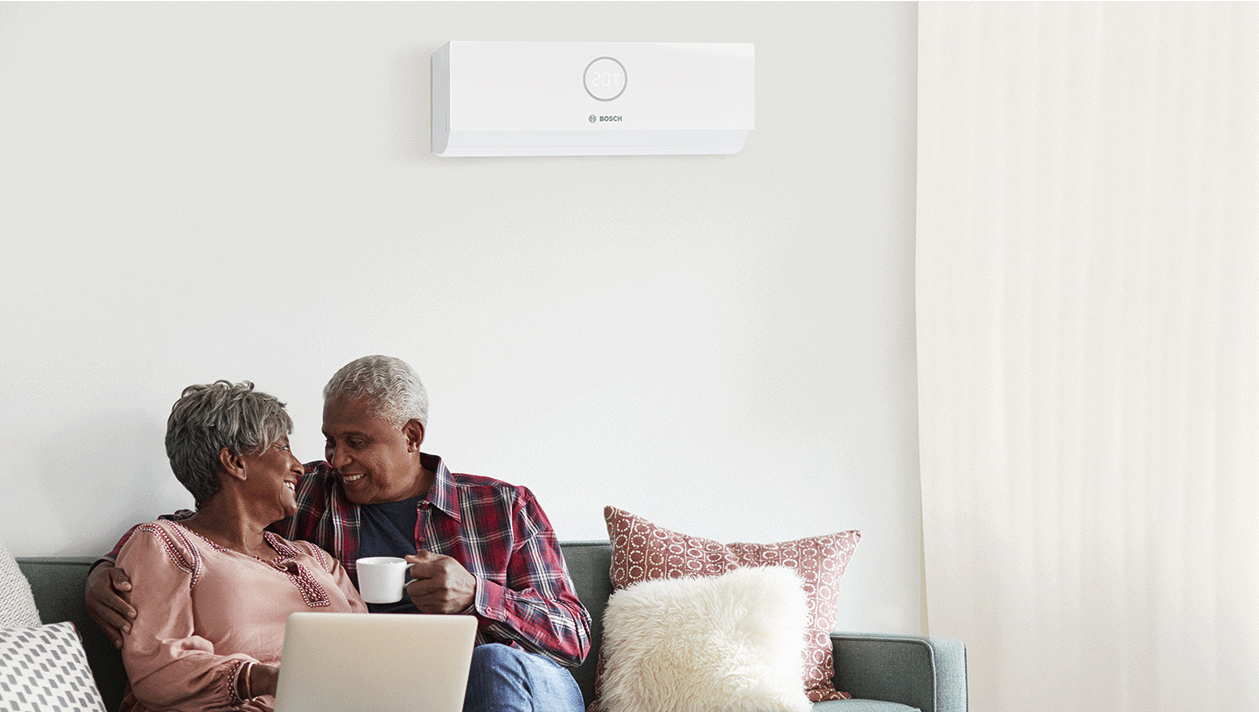 Ein &auml;lteres Ehepaar sitzt auf einem Sofa und l&auml;chelt sich an. Der Mann h&auml;lt eine Tasse in der Hand, und auf dem Scho&szlig; der Frau steht ein Laptop. An der Wand &uuml;ber ihnen ist eine wei&szlig;e Bosch-Klimaanlage angebracht.