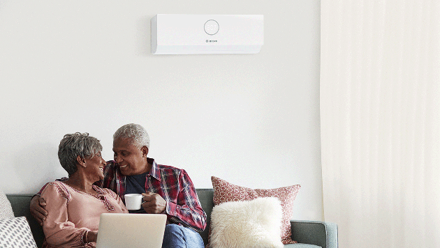 Ein &auml;lteres Ehepaar sitzt auf einem Sofa und l&auml;chelt sich an; daneben stehen ein Laptop und Kissen. An der Wand hinter ihnen ist eine wei&szlig;e Klimaanlage angebracht, die vom Bosch Werkskundendienst fachgerecht gewartet wird.